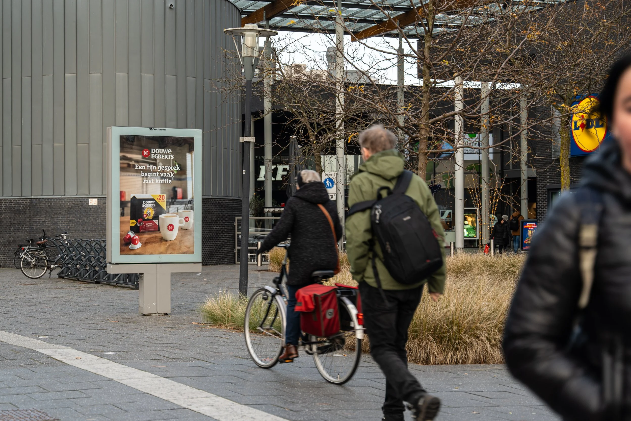 Mensen lopen en fietsen buiten een winkelcentrum met een reclamebord voor Douwe Egberts koffie.