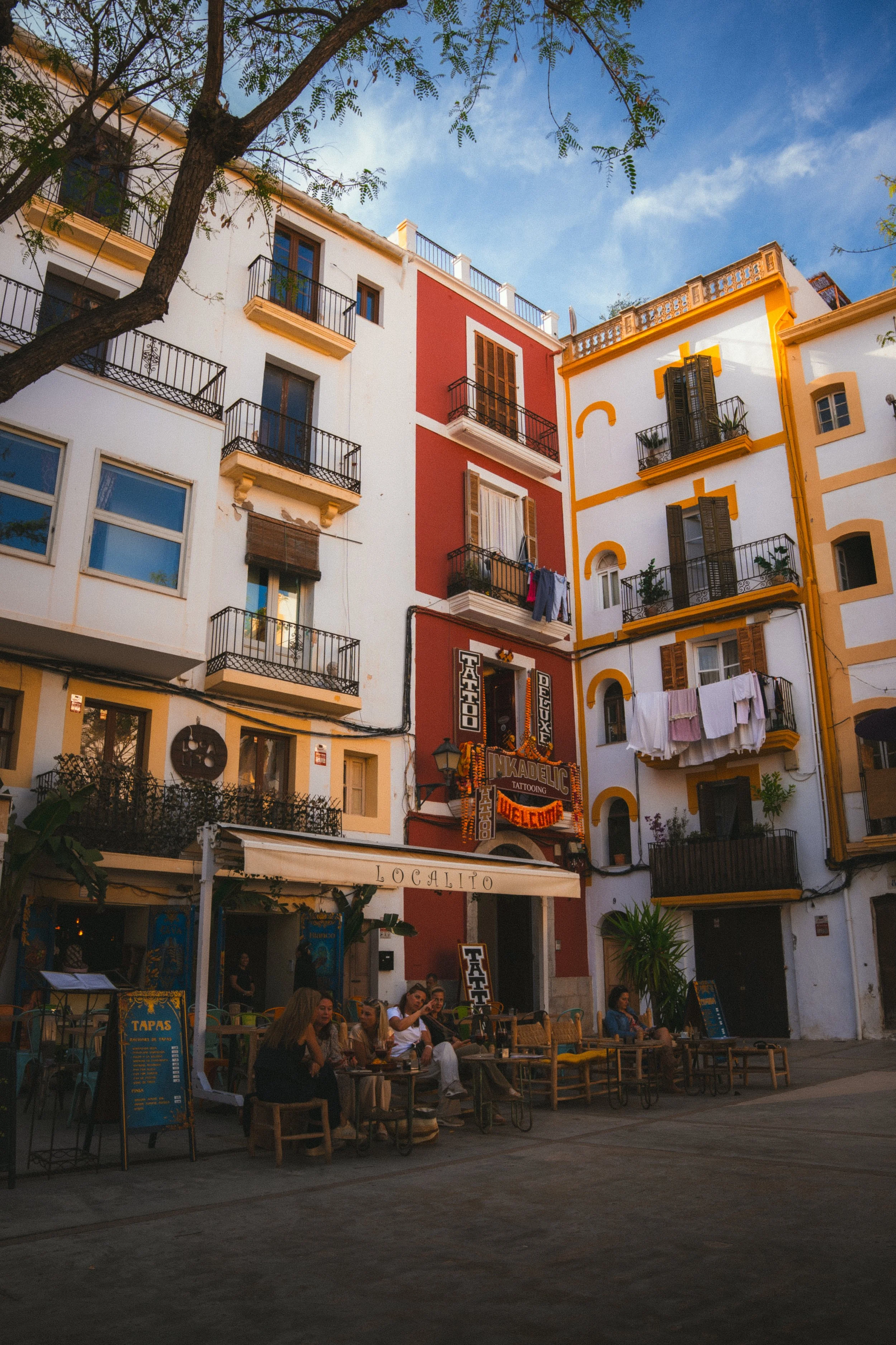 Gezellig straatbeeld met mensen zitten buiten een tapasrestaurant, omringd door kleurrijke gebouwen met balkons en hangende was. Blauwe lucht en een boom tak boven.