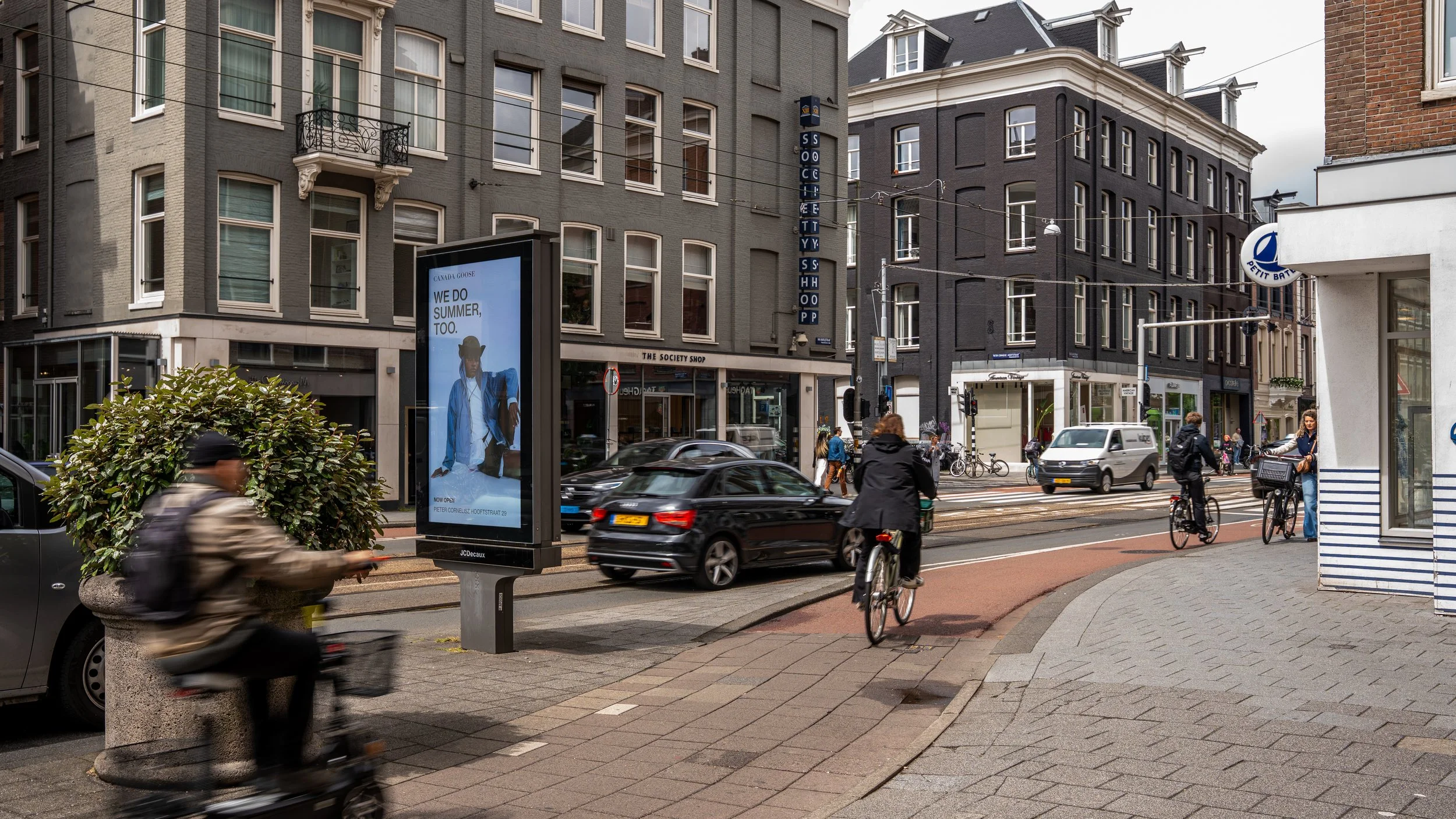 Straatbeeld met mensen op fietsen en wandelend, auto's, verkeerslichten, digitale folder als reclame, diverse gebouwen in historische stijl, een bushalte en tramlijnen in een stedelijke omgeving.