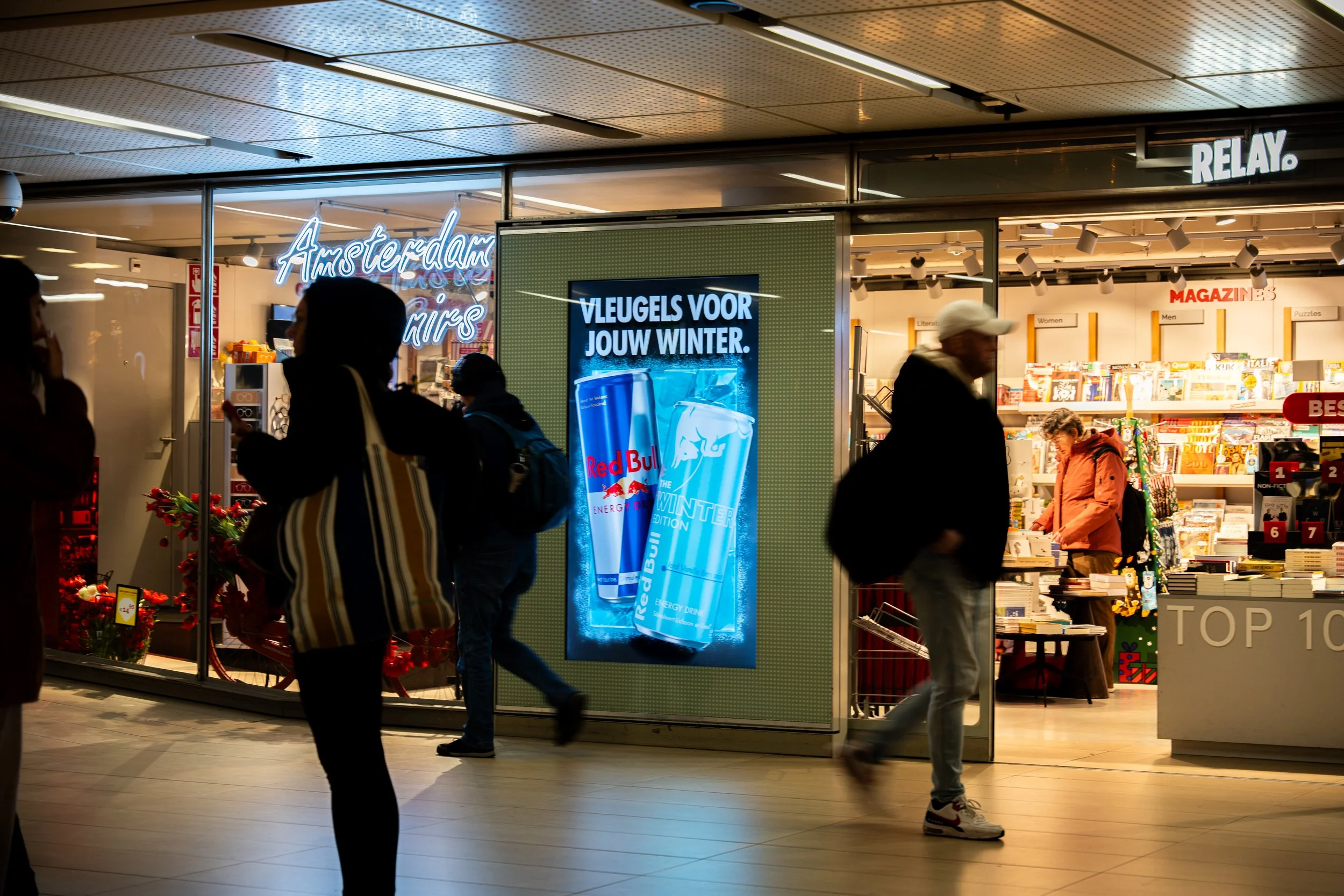 Binnen- en buitendetails van een winkel in Nederland. Op het digitale bord staat 'VLEUGELS VOOR JOUW WINTER' met Red Bull energiedrankjes. Mensen lopen voorbij de winkel, met enkele shoppers binnen, op een avond of daglicht na zonsondergang.