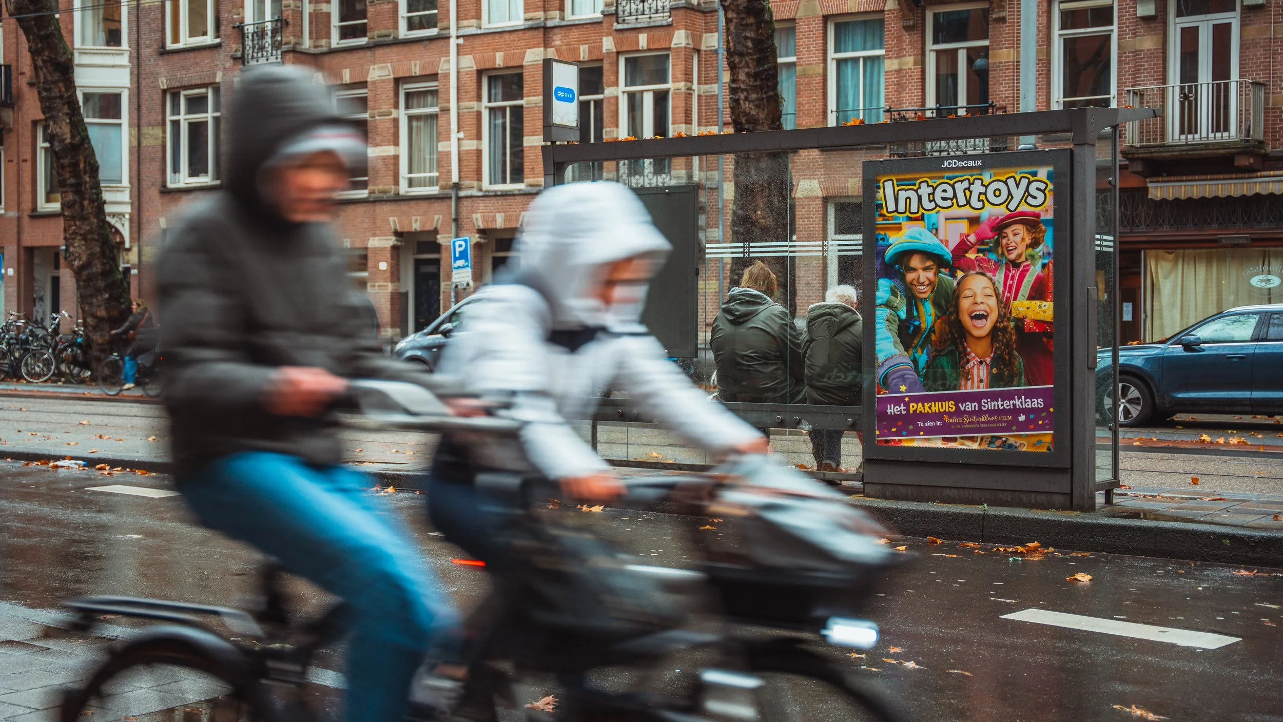 Twee mensen rijden op een scooter over een regenachtige straat met een bushalte en een postershop op de achtergrond.