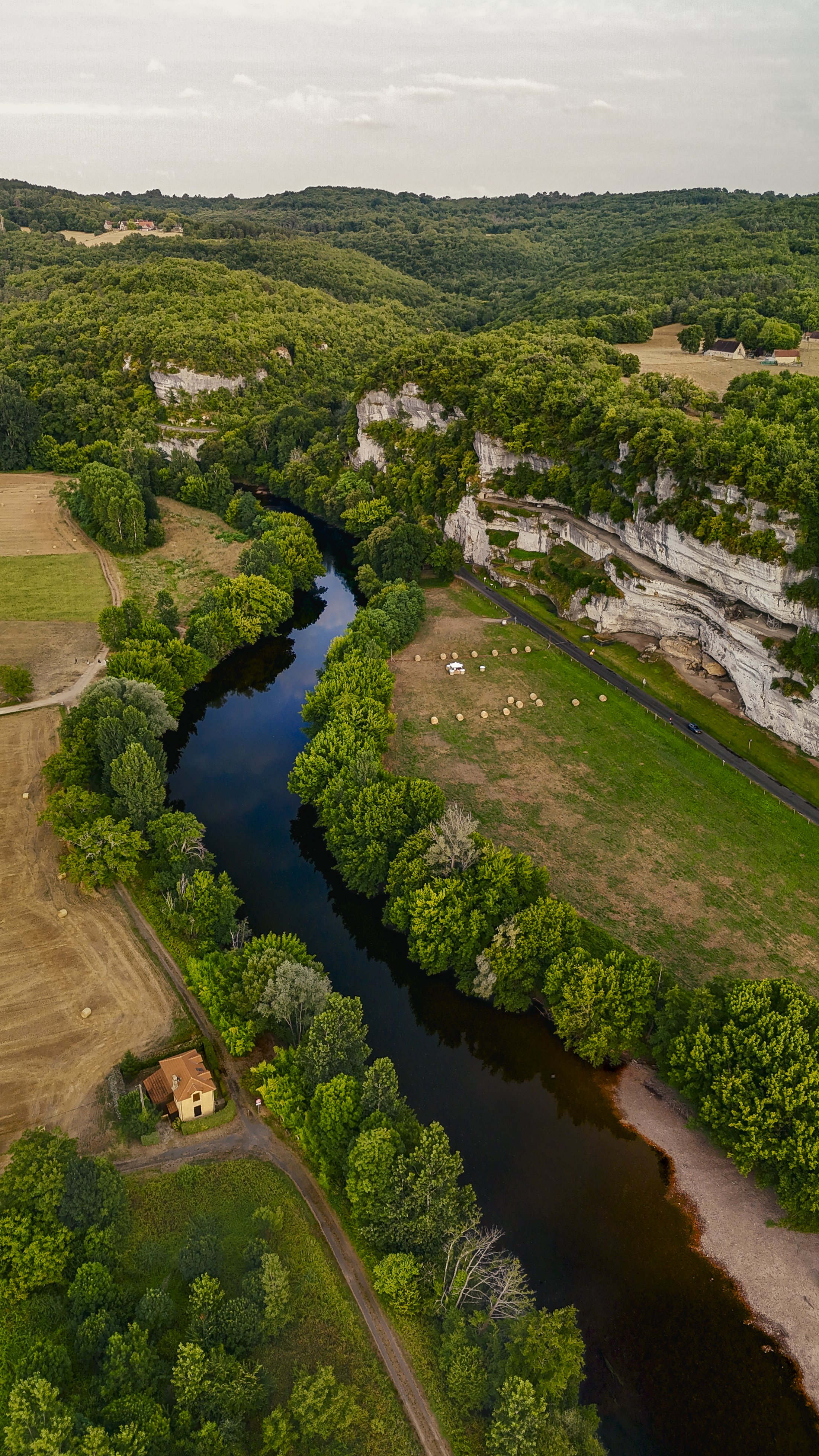 Luchtfoto van een rivier die door groene velden en bossen stroomt, met witte kalksteen kliffen langs de rechterkant en een klein huis links aan de rand van het bos.
