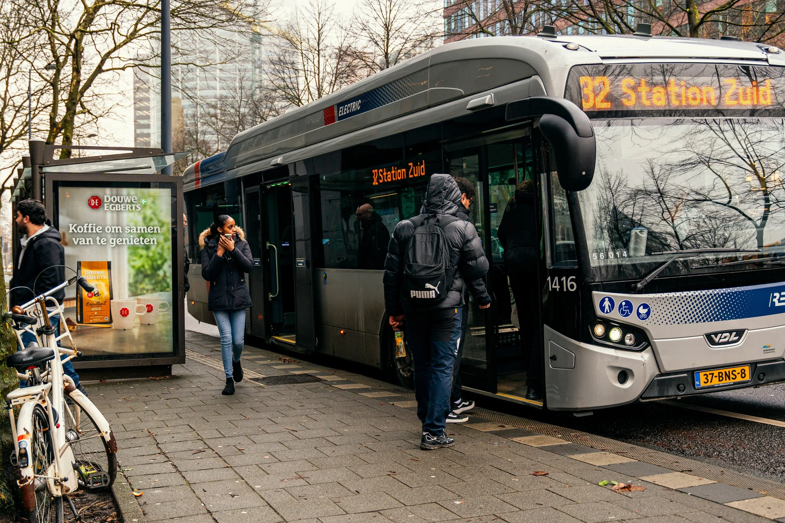 Een stadsbus staat klaar om passagiers te laten instappen op een bushalte in Nederland, met mensen die wachten en in- en uitstappen op een druilerige dag.