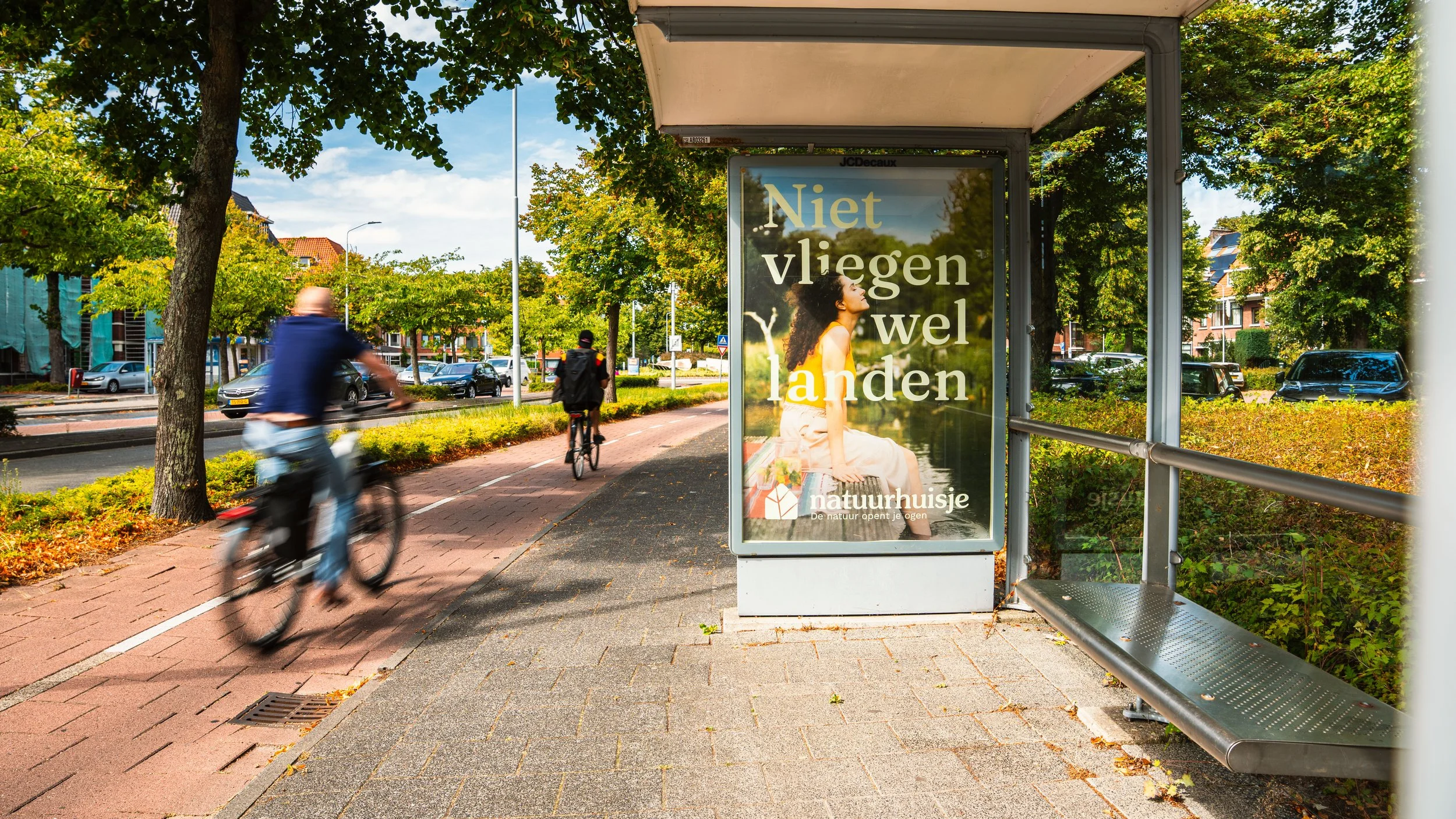een bushokje met een advertentie die zegt 'Niet vliegen wel landen' met een vrouw die in de natuur kijkt, op een zonnige dag met mensen die fietsen voorbijrijden