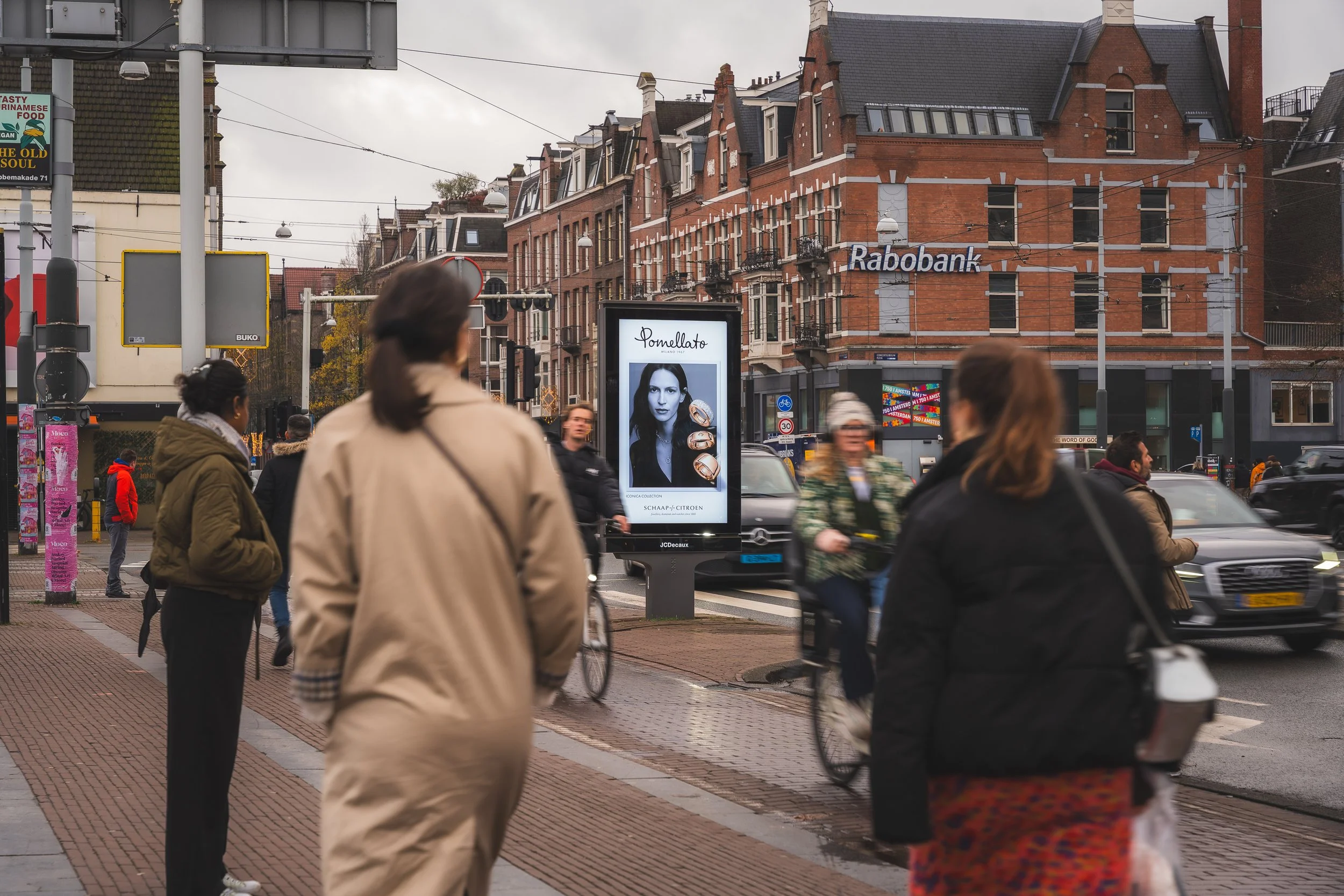 Stadsplein met mensen die wachten en fietsen, reclamebord met een vrouw en sieraden, achtergrond met gebouwen en winkel 'Rabobank'.