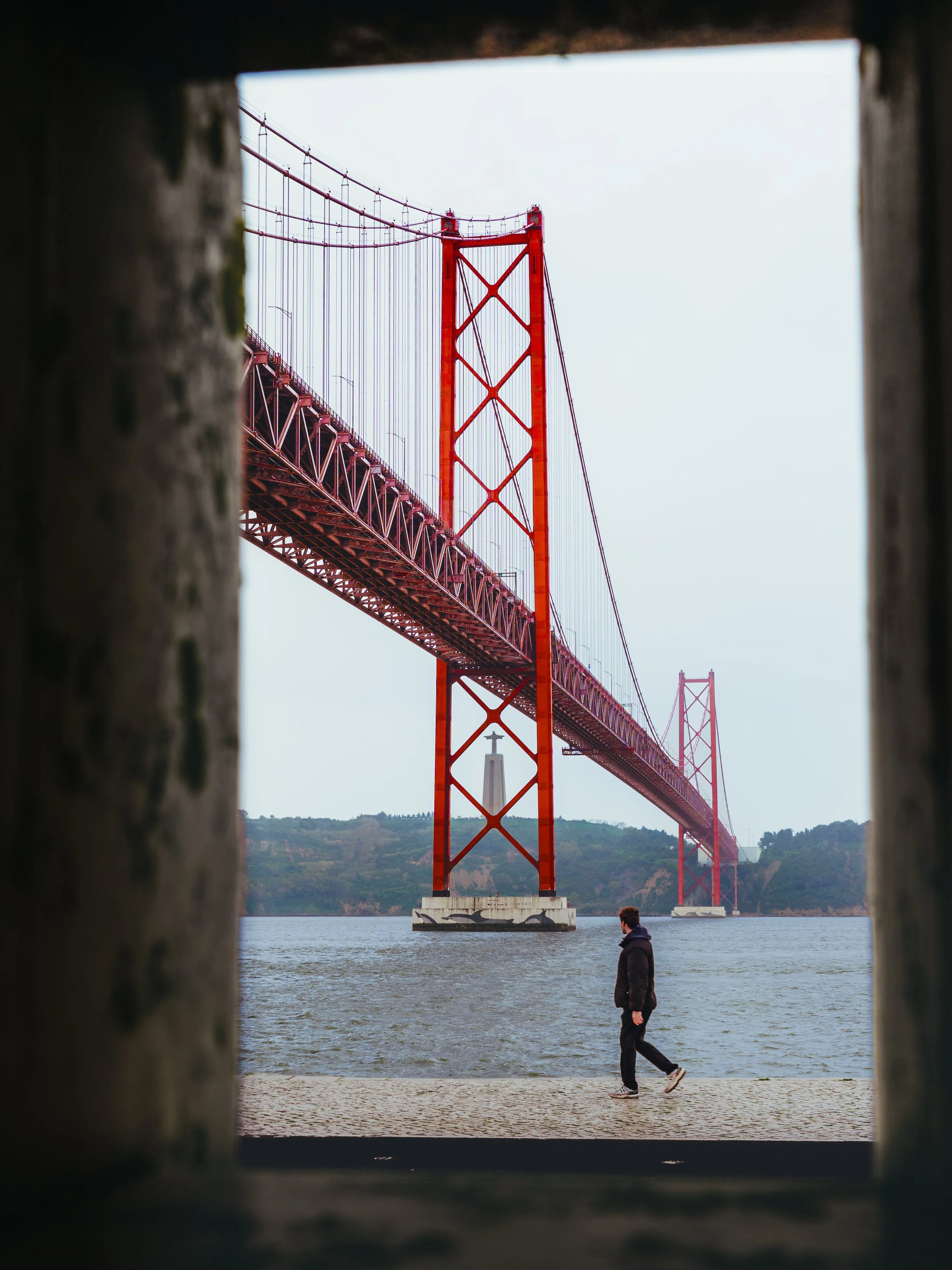 Kijk door een raam uit hout naar de Ponte 25 de Abril-brug in Lissabon met een wandelaar op de promenade.