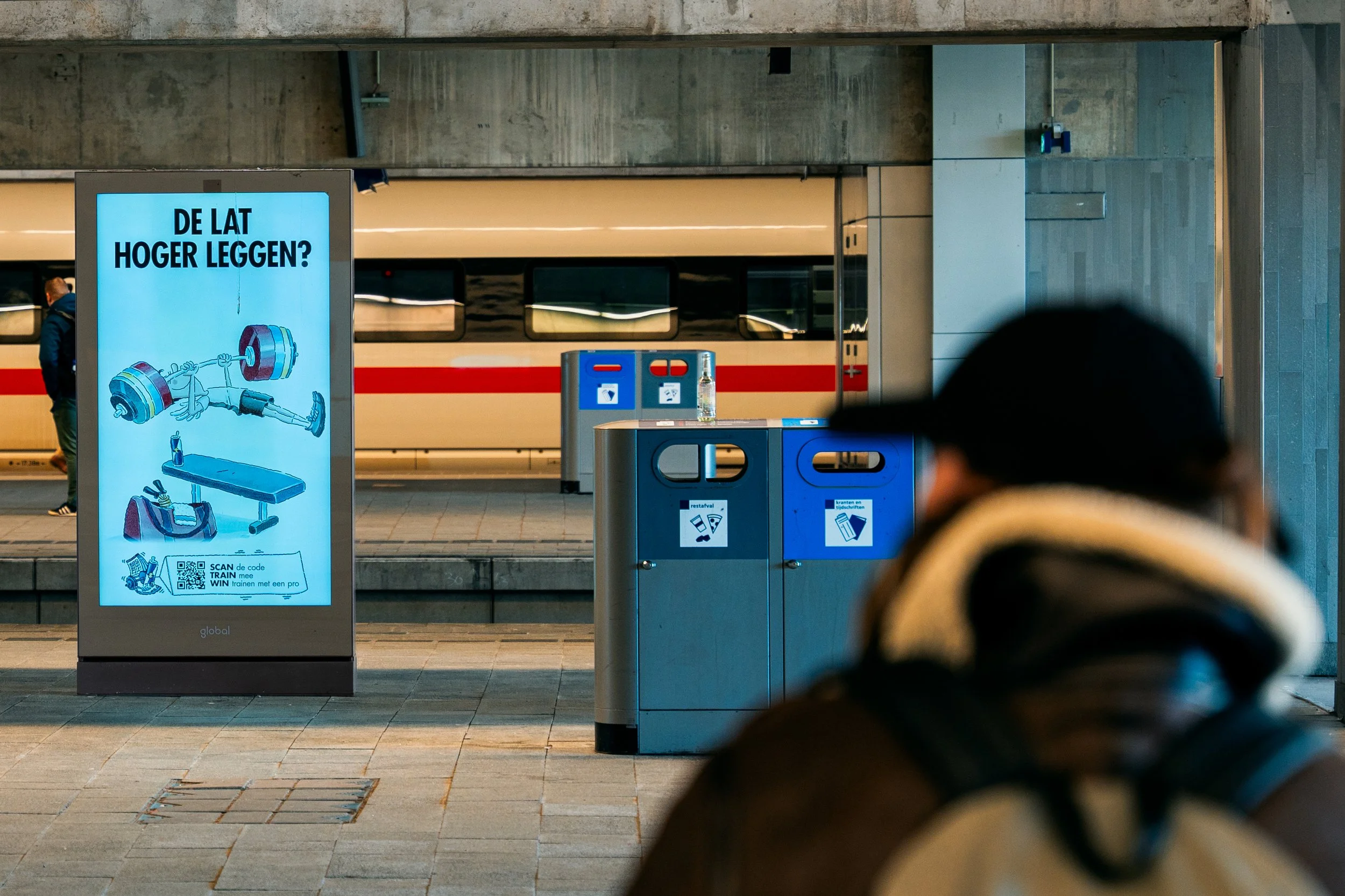 Een persoon met een zwarte hoed en beige sjaal wacht in een treinhalte. Op de achtergrond is een trein te zien en een digitale informatiescherm met een afbeelding van sportschoolapparatuur en tekst in het Nederlands.