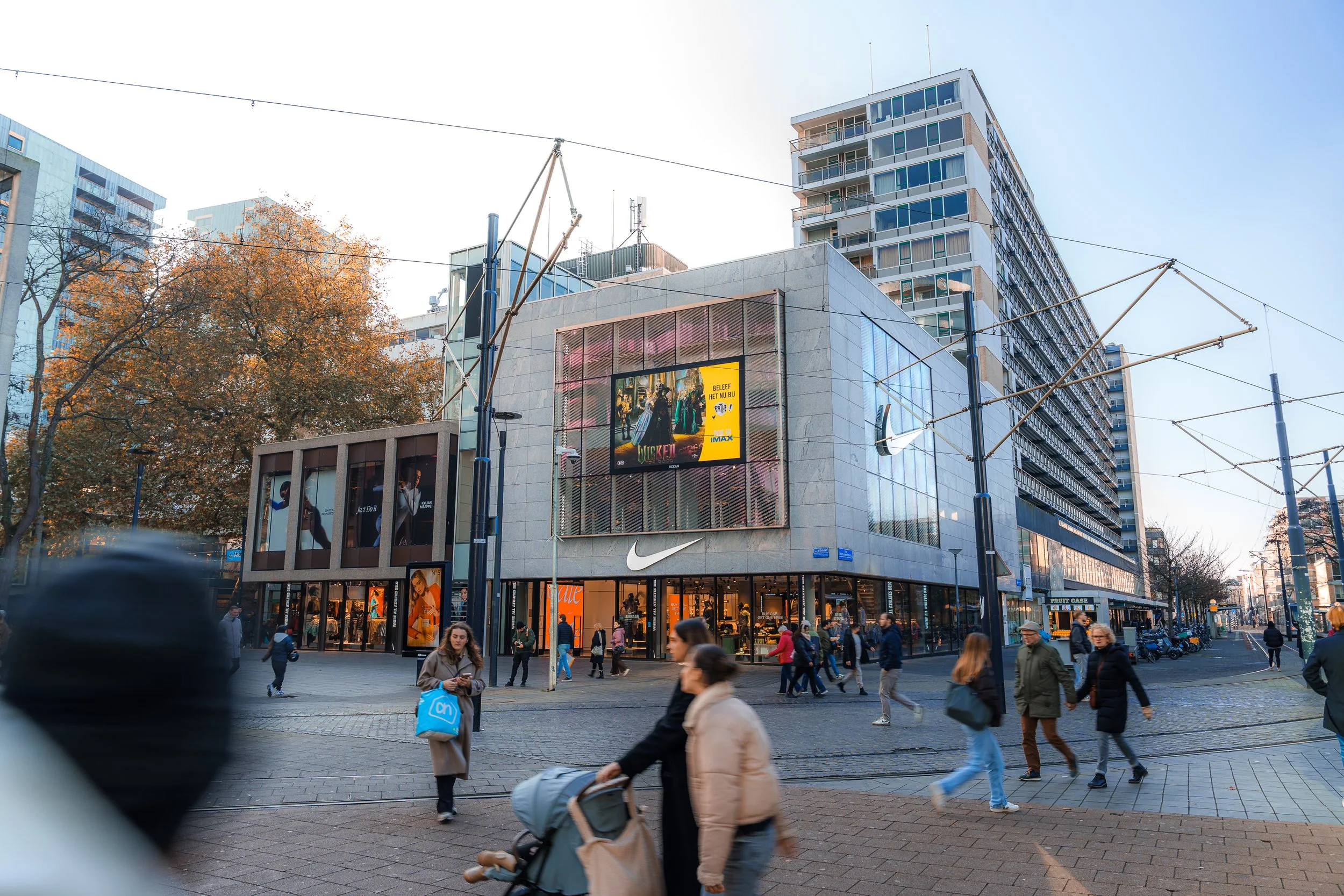 Een drukke stadsplein met winkels, waaronder een Nike winkel, en een groot digital billboard op een modern gebouw. Mensen lopen over het plein, enkele met kinderwagens en boodschappentassen, en er staan vooral bomen met herfstbladeren.