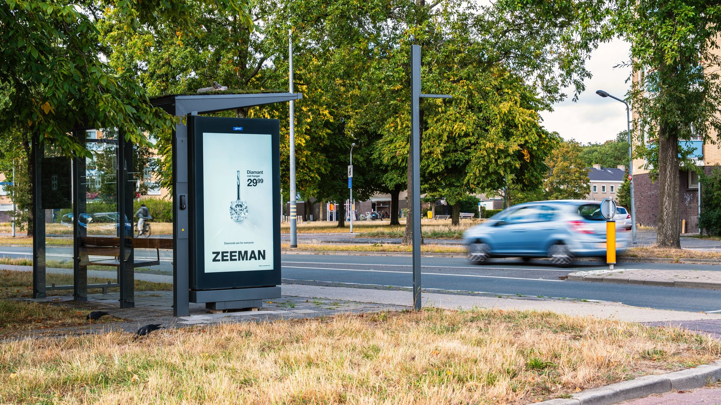 Een bushokje met een reklamebord langs een straat met een rijdende auto, bomen en een lantaarnpaal op de achtergrond.