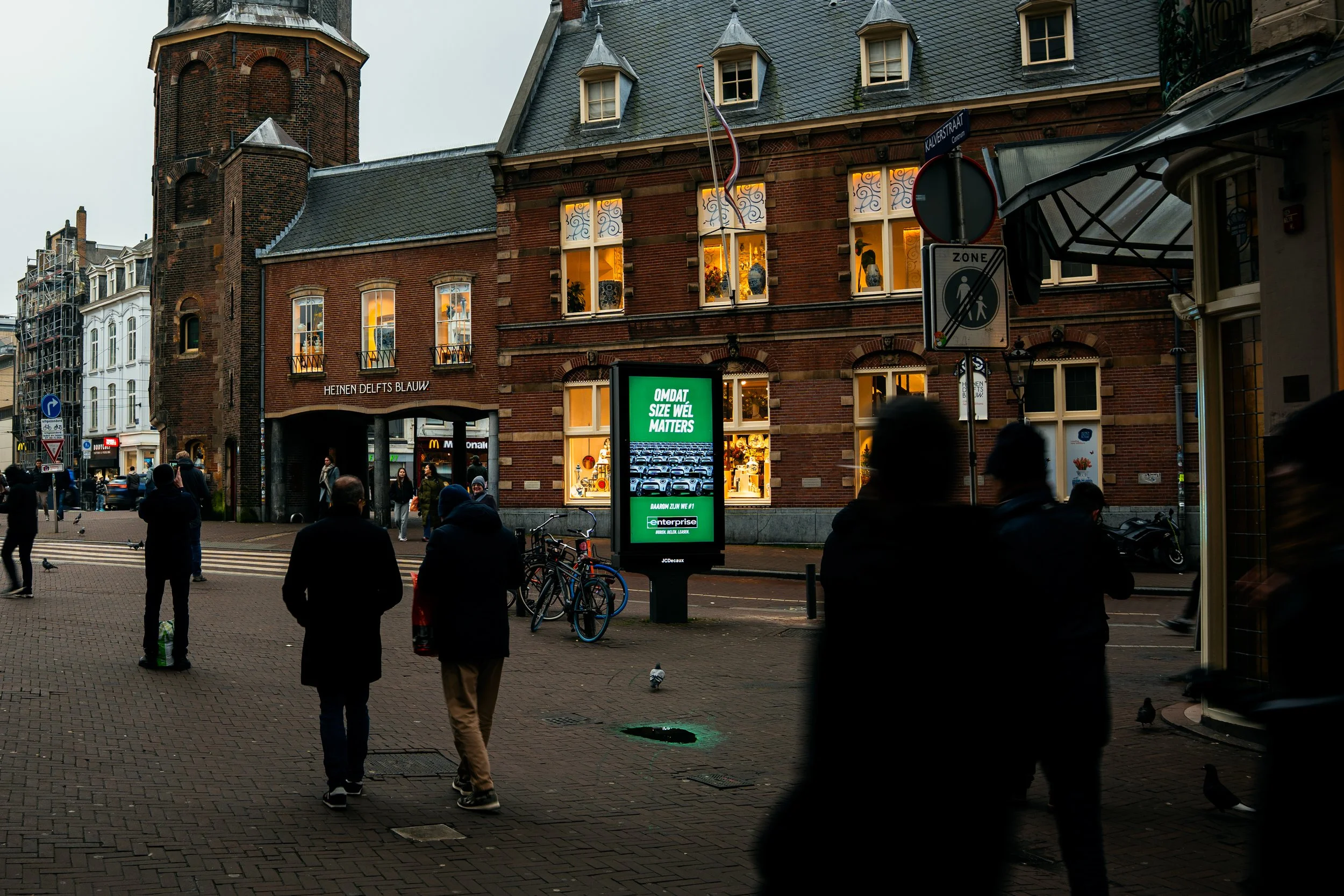 Straatbeeld met mensen, fietsen, een digitaal billboard en oude gebouwen in de avond. 