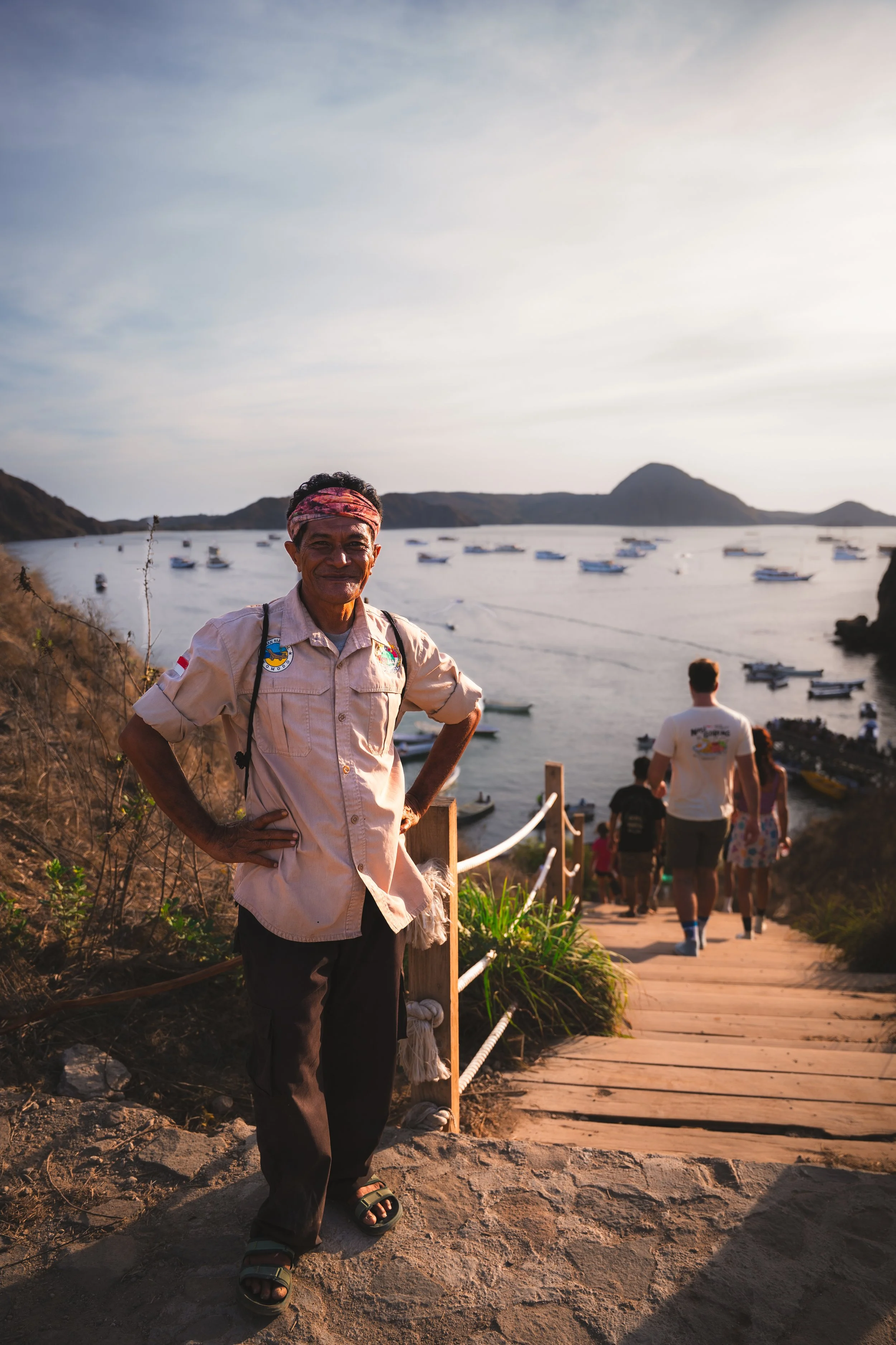 Vrouw staat bij ingang van een trap die naar een haven met boten leidt, met een berg en een blauwe lucht op de achtergrond, tijdens zonsondergang.