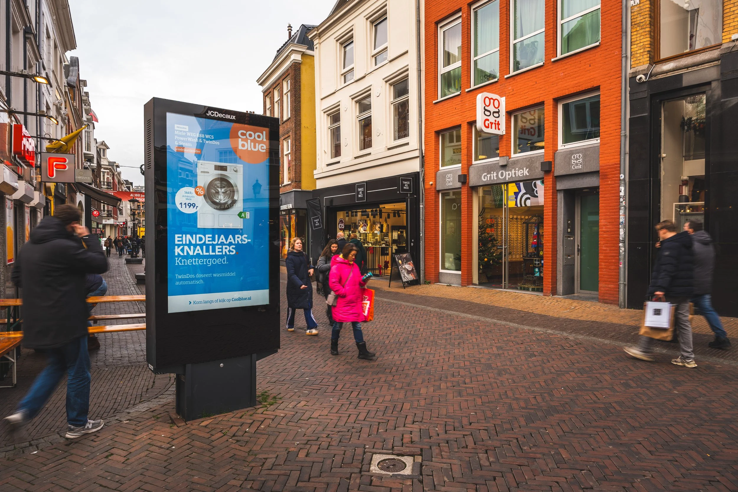 Een drukke winkelstraat met mensen die lopen en winkelen. Er staat een groot digitaal reclamebord dat een wasmachine adverteert, en er zijn winkels zoals Grit Optiek en een ander met een zwart gevel. De straat is geplaveid met houten stenen en er han