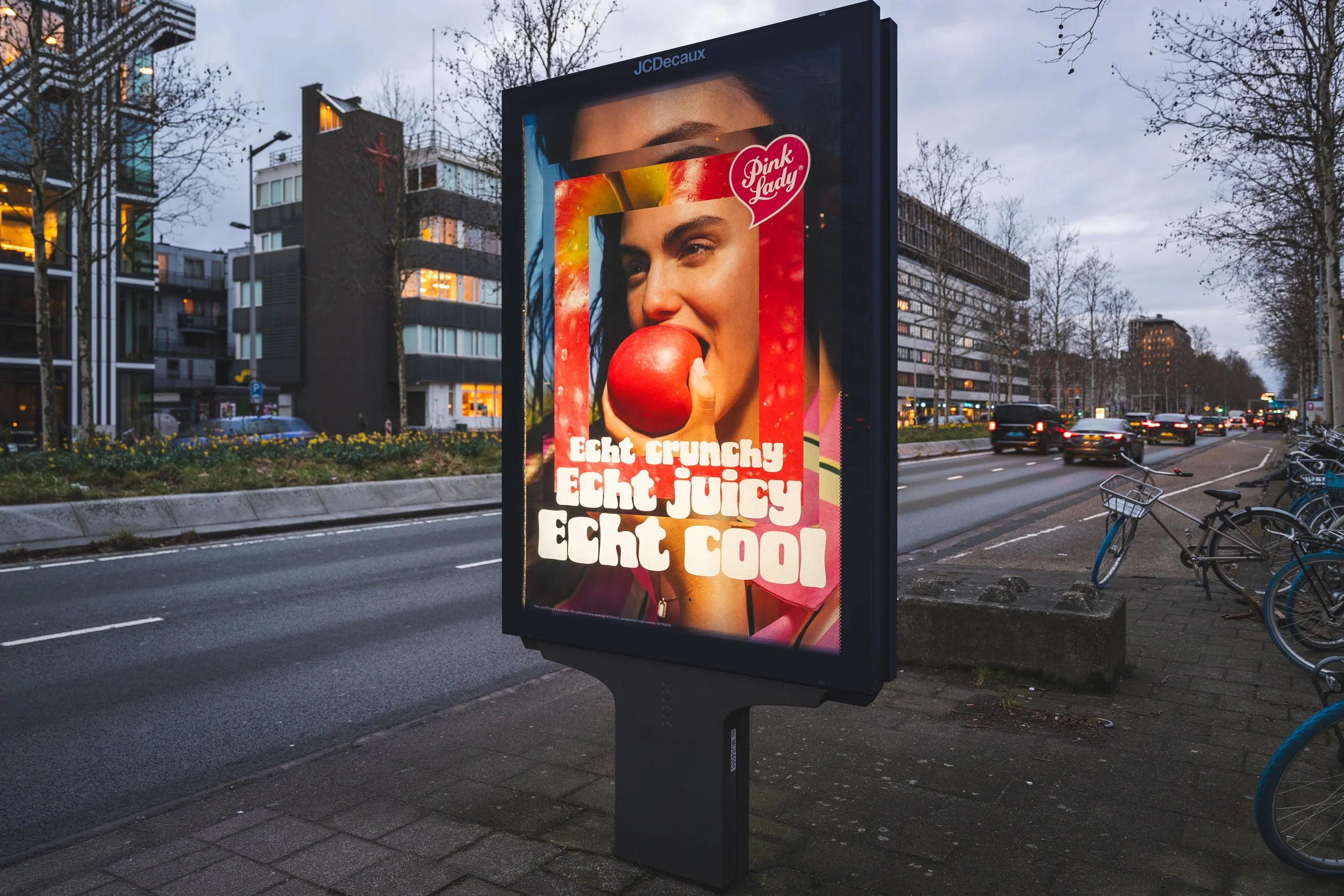 Een digitale billboard langs een straat in de avond met een advertentie van een vrouw die een rode appel houdt en er aan bijt. De advertentie bevat teksten in het Nederlands en Engels: 'Echt crunchy', 'Echt juicy', 'Echt cool'.