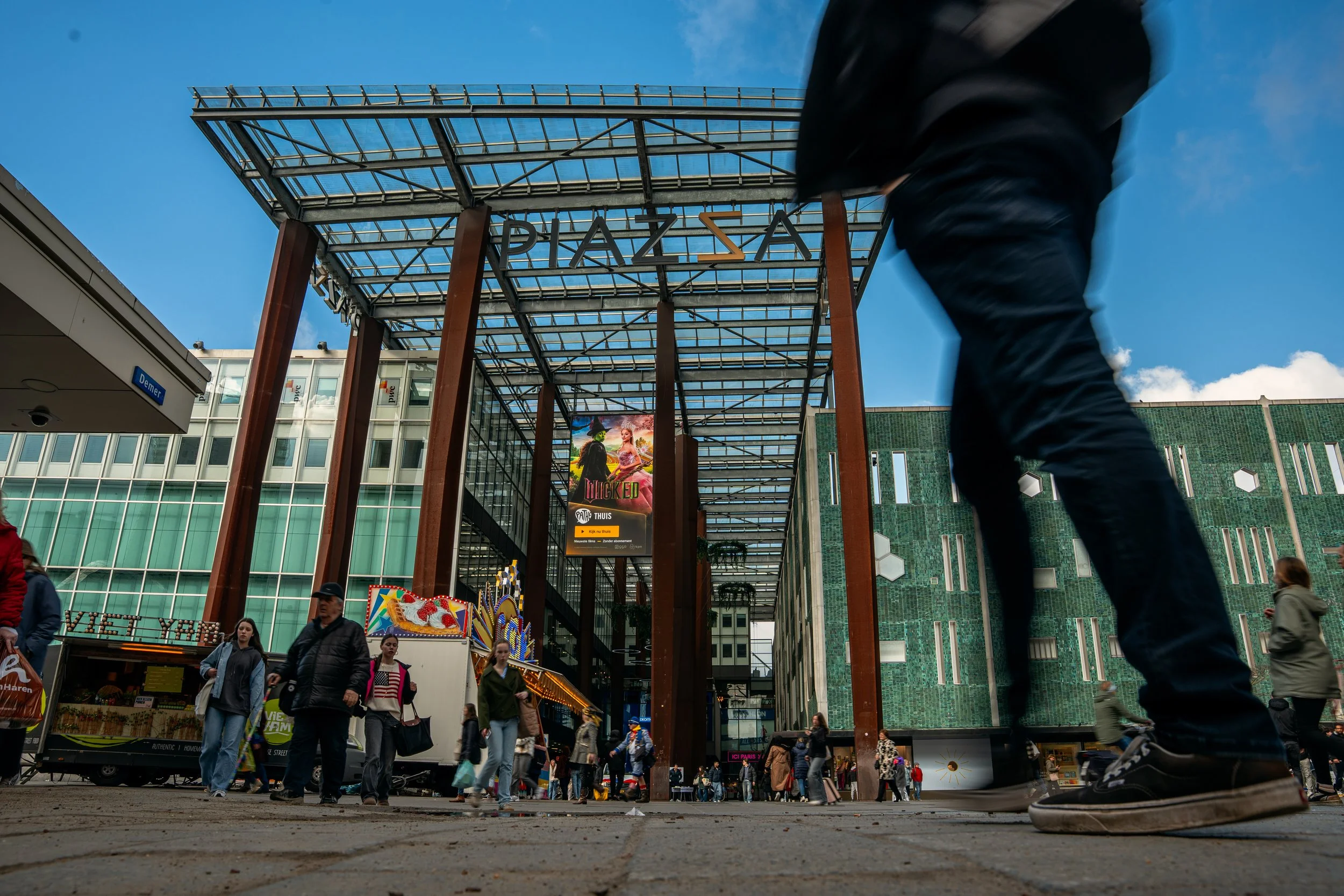Mensen wandelen bij een modern winkelcentrum met een glazen en metalen ingang, terwijl een persoon op de voorgrond snel loopt.