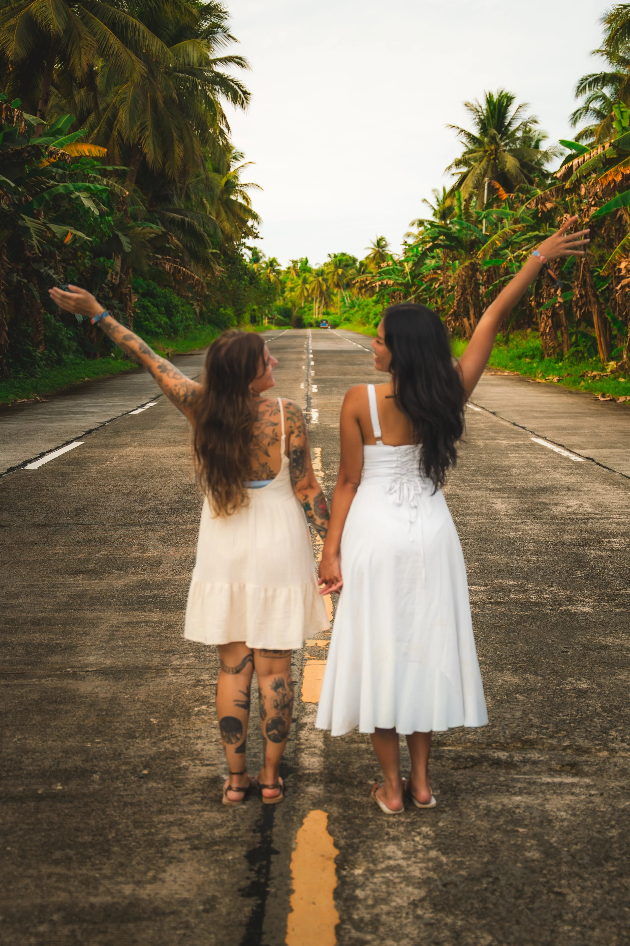Twee vrouwen lopen hand in hand op een lege weg omringd door palm- en bananenbomen, beiden met uitgestrekte armen.