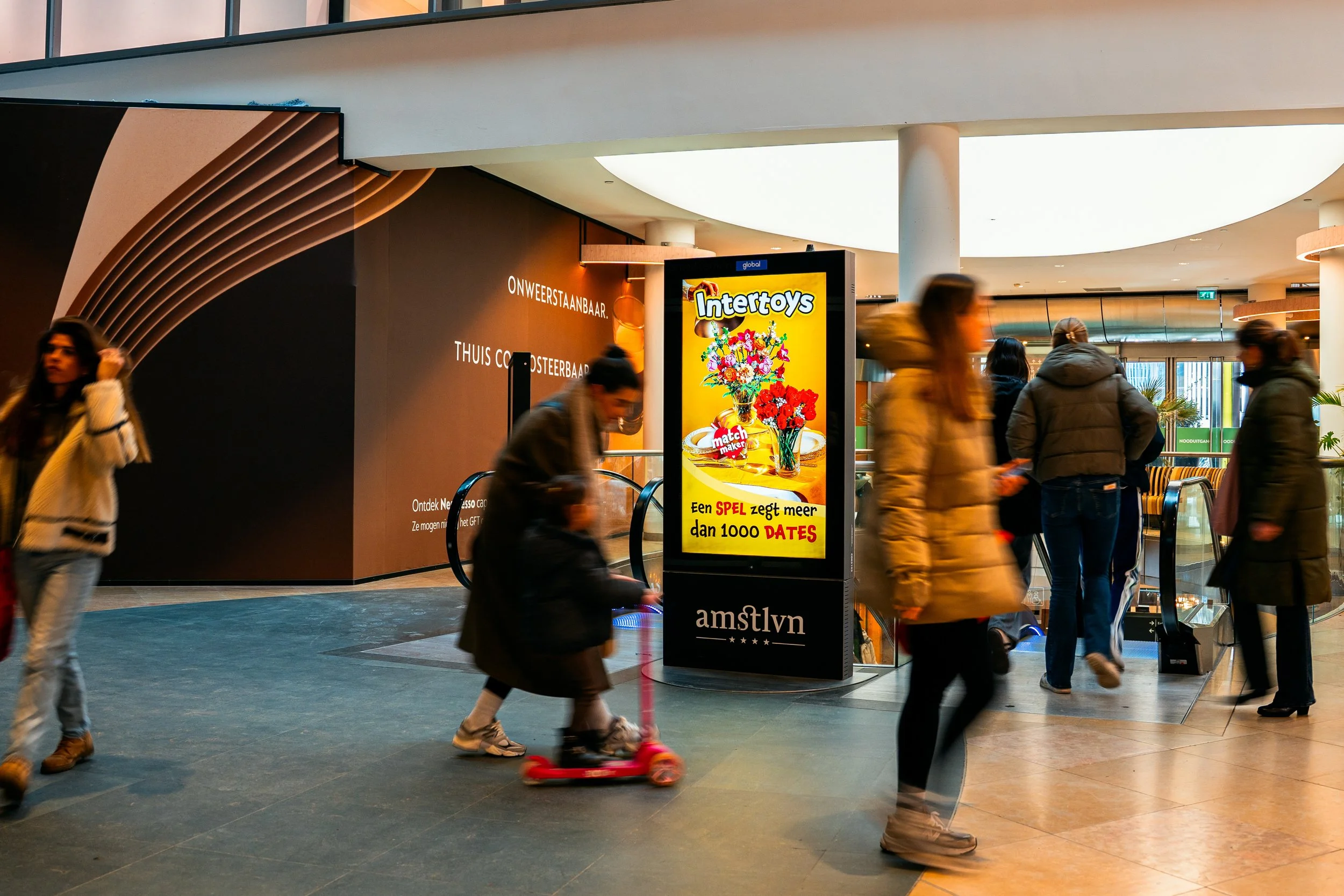 Binnen in een winkelcentrum staan meerdere mensen, waaronder een kind op een roze scooter. In het midden staat een digitale reclamezuil met een advertentie voor Intertoys. Op de achtergrond zijn roltrappen en een muur met teksten in het Nederlands te