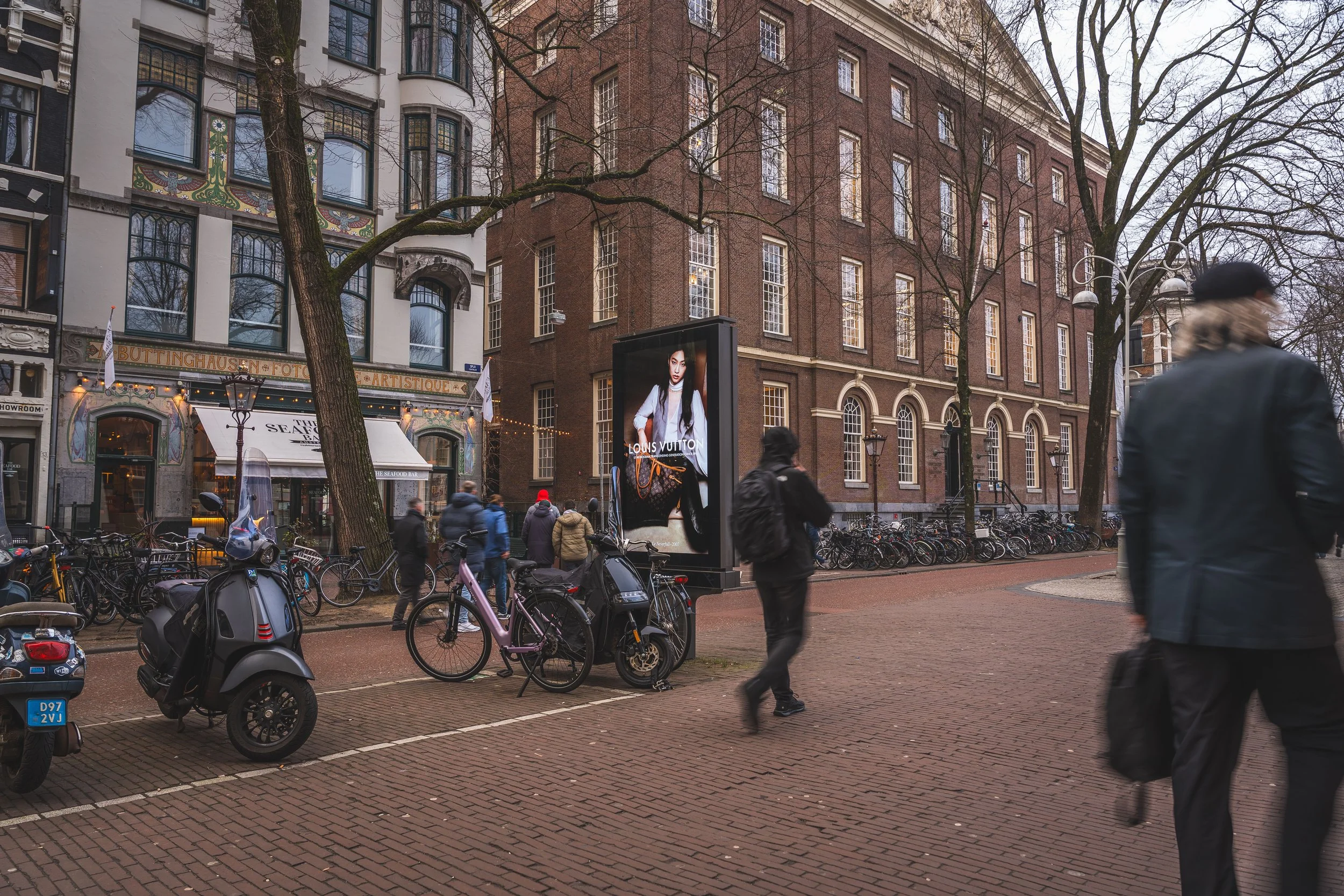 Stadsplein met fietsen, scooters en voetgangers. Een advertentiescherm voor Louis Vuitton, oude gebouwen en bomen op de achtergrond.