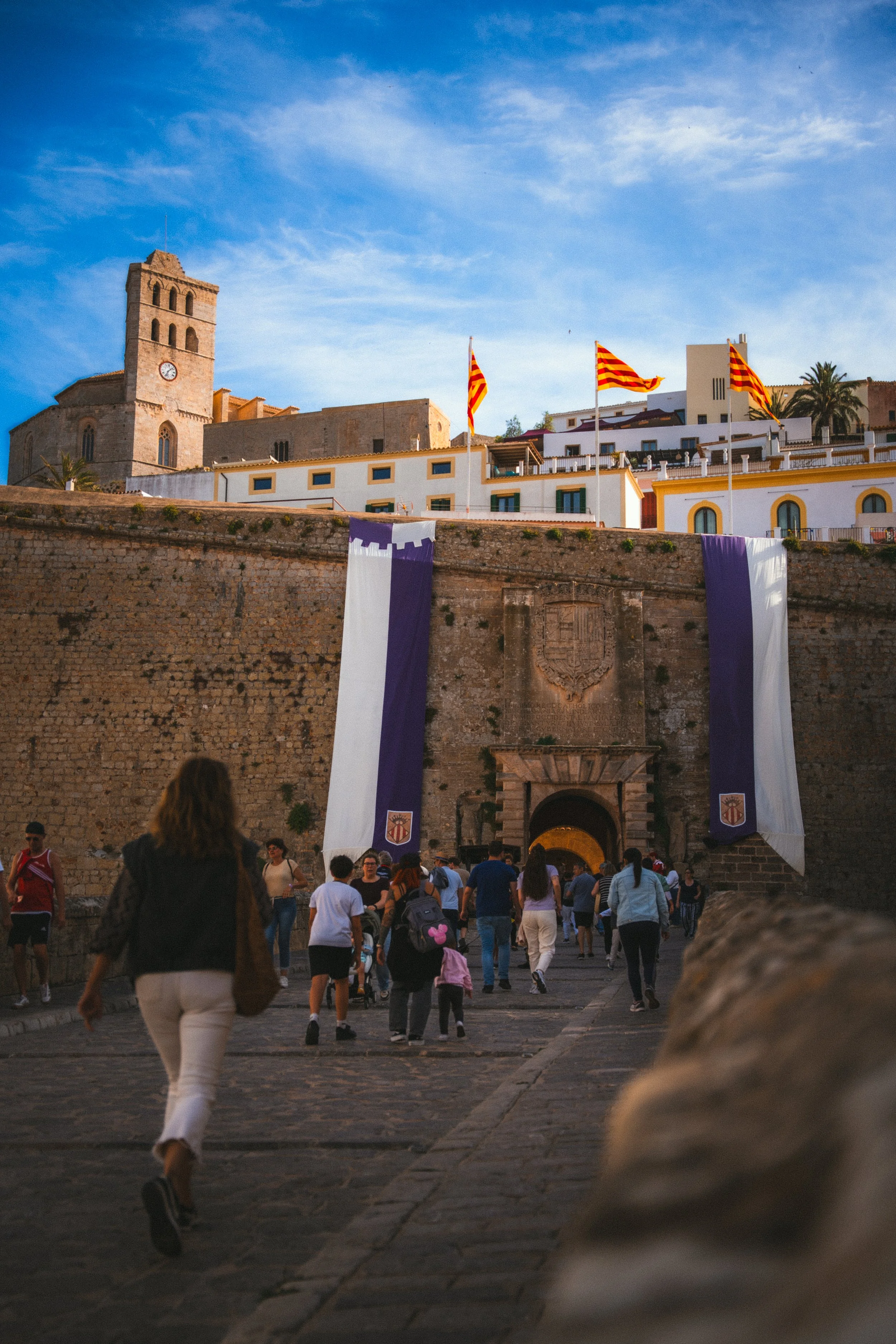 Mensen lopen onder een oude stadsmuur met vlaggen en een kerk op de achtergrond, tijdens een zonnige dag.