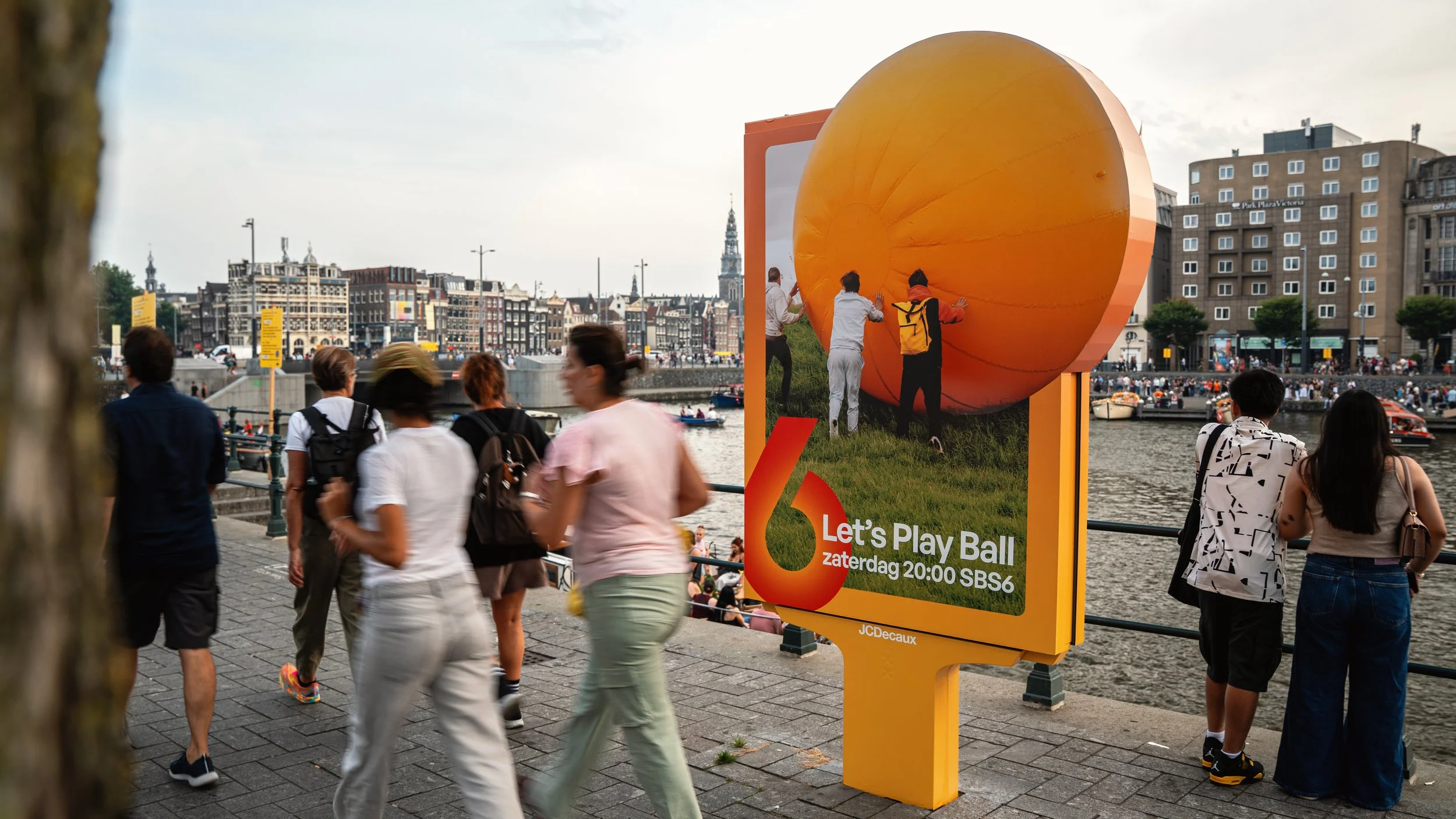 Mensen wandelen langs een rivierkant met grote poster die een ballon en mensen toont, in een stad met veel gebouwen op de achtergrond.