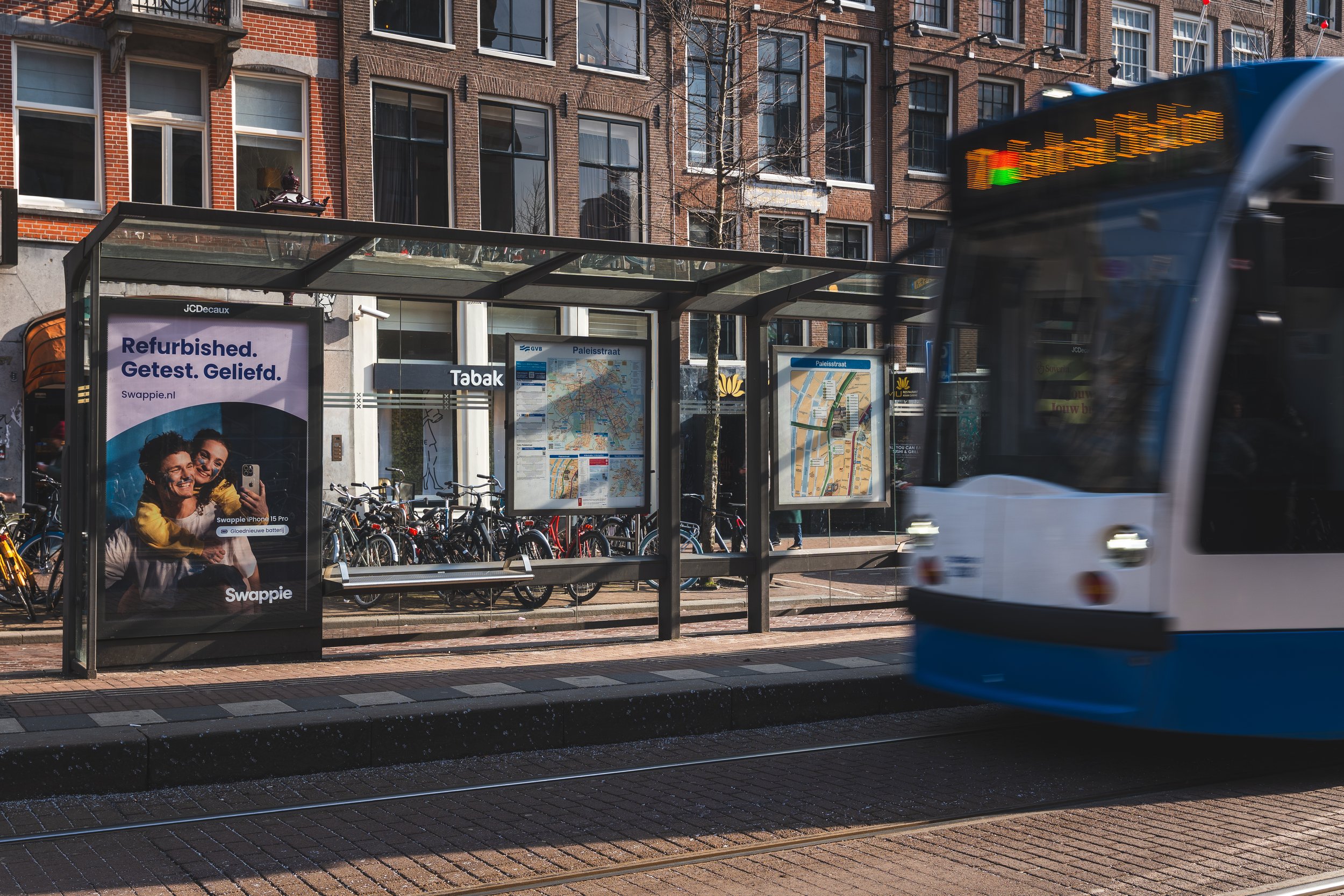 Een tram komt aan bij een bushalte in een stadswijk met bakstenen gebouwen en fietsen. Op de bushalte bevinden zich posters en een routekaart.