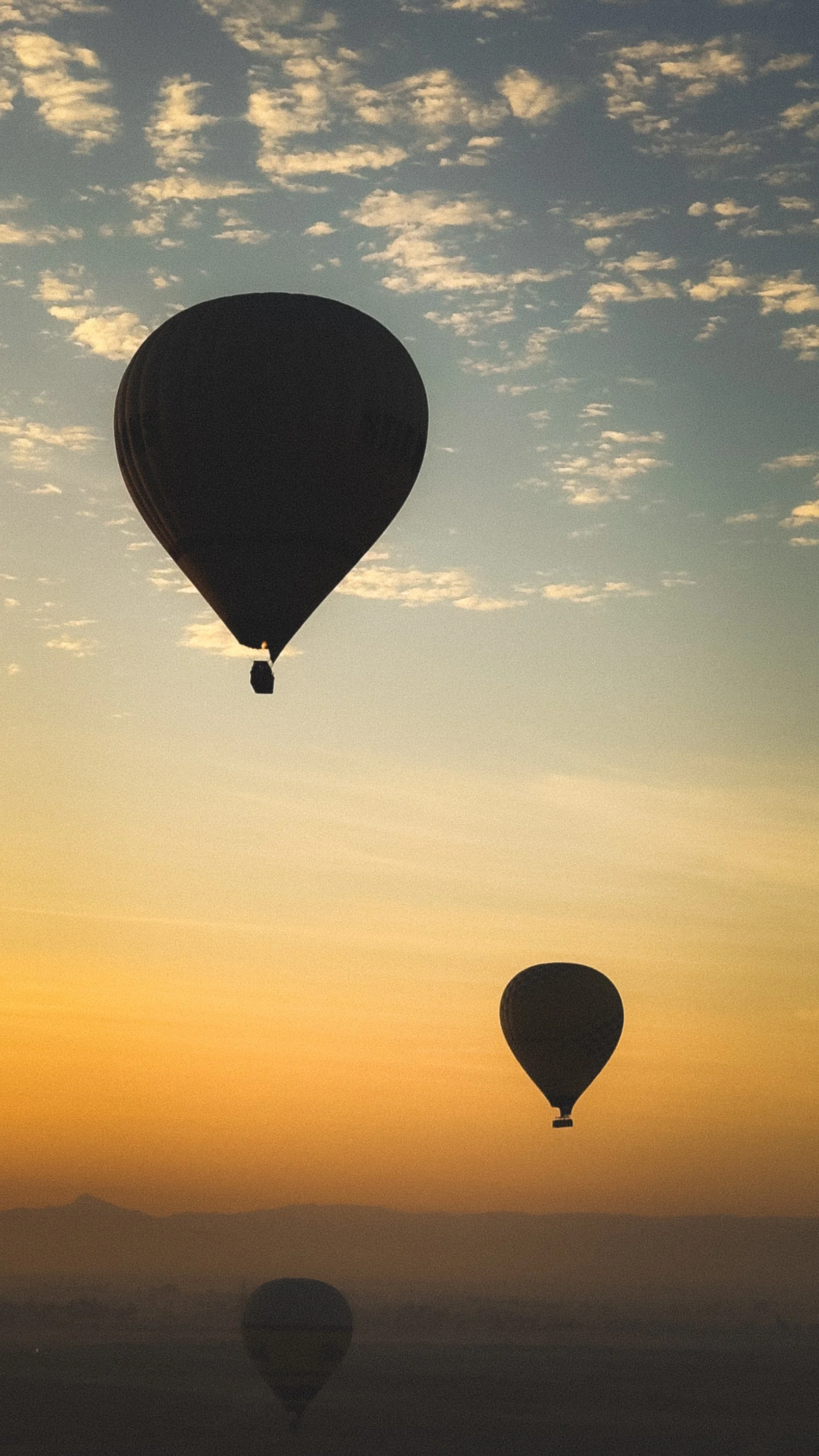 Twee hete luchtballonnen zweven boven een landschap bij zonsopgang of zonsondergang, met een heldere hemel en enkele wolken.