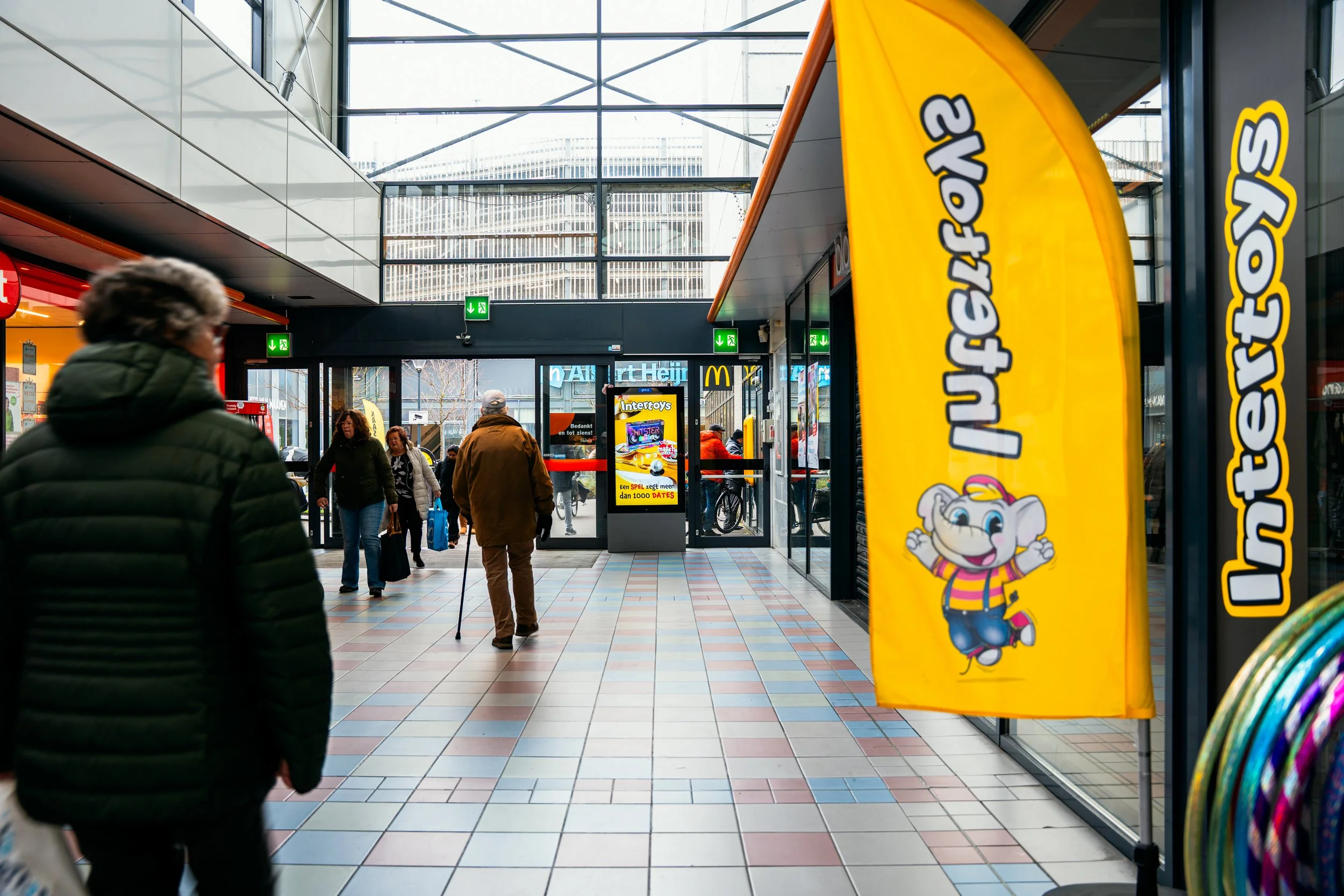 Binnenruimte van een winkelcentrum met mensen die naar de uitgang lopen, een vlag met een tekenfilmkoala en een scherm met reclame voor Intertoys.