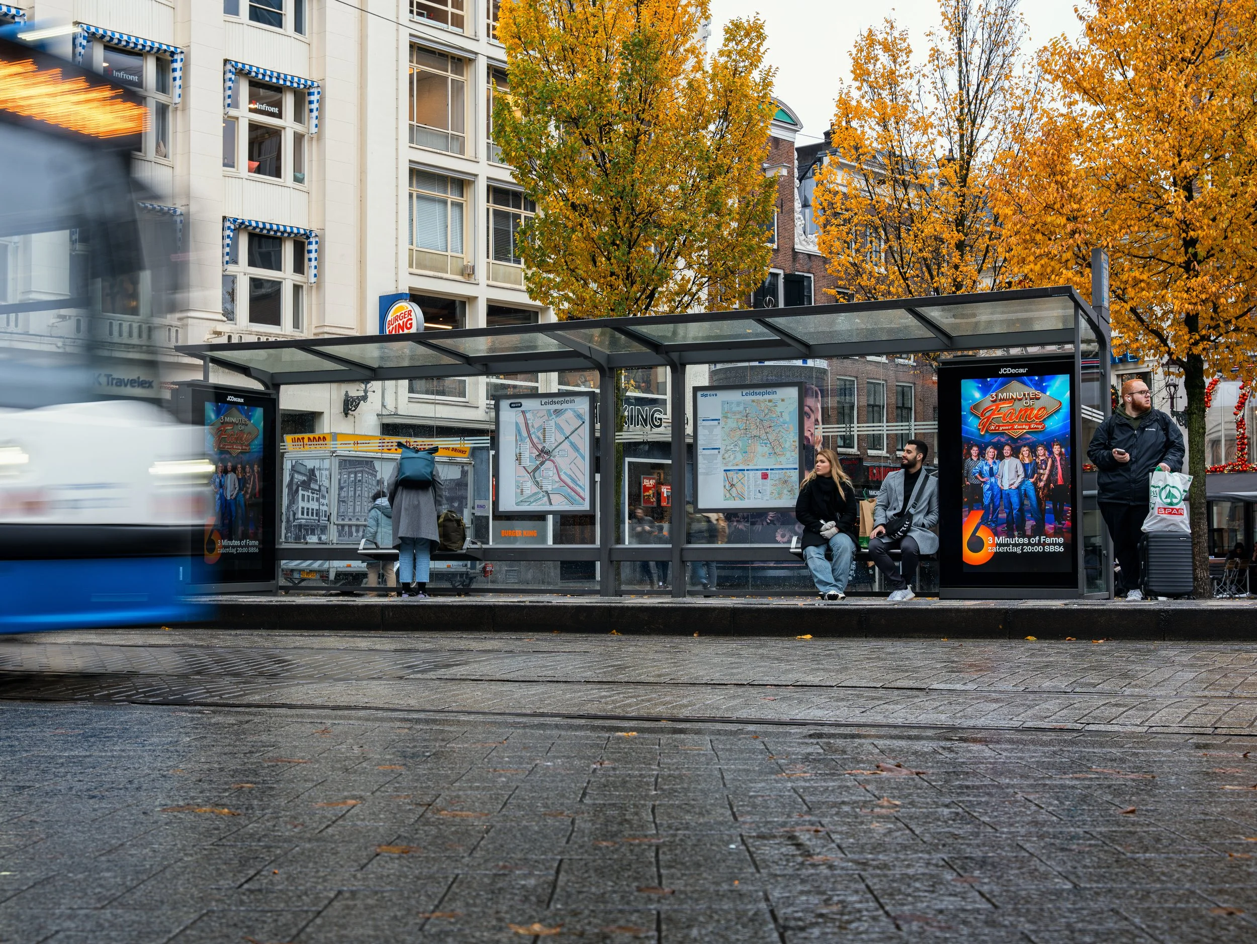 Een bushalte op straat in de herfst met mensen die wachten en een tram die langsrijdt, omgeven door bomen met oranje bladeren.