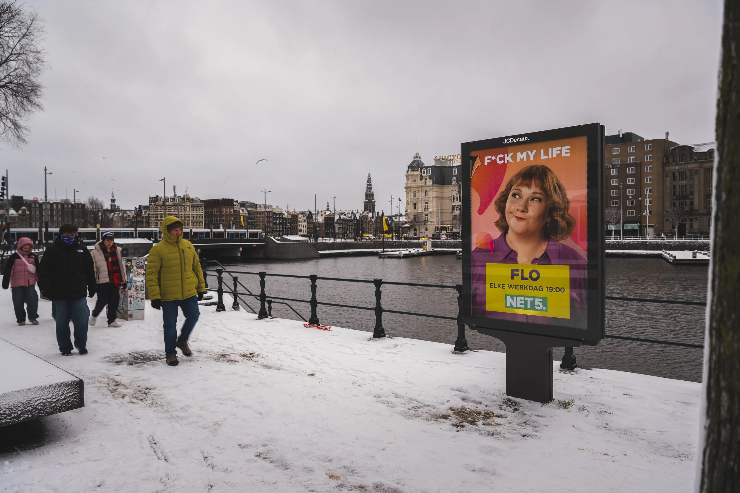 Een winterse rivierkant in Amsterdam met een digitale advertentiebord dat een vrouw afbeeldt en teksten bevat, waaronder 'F*CK MY LIFE', 'FLO', 'ELKE WERKDAG 19:00', en 'NET5'.