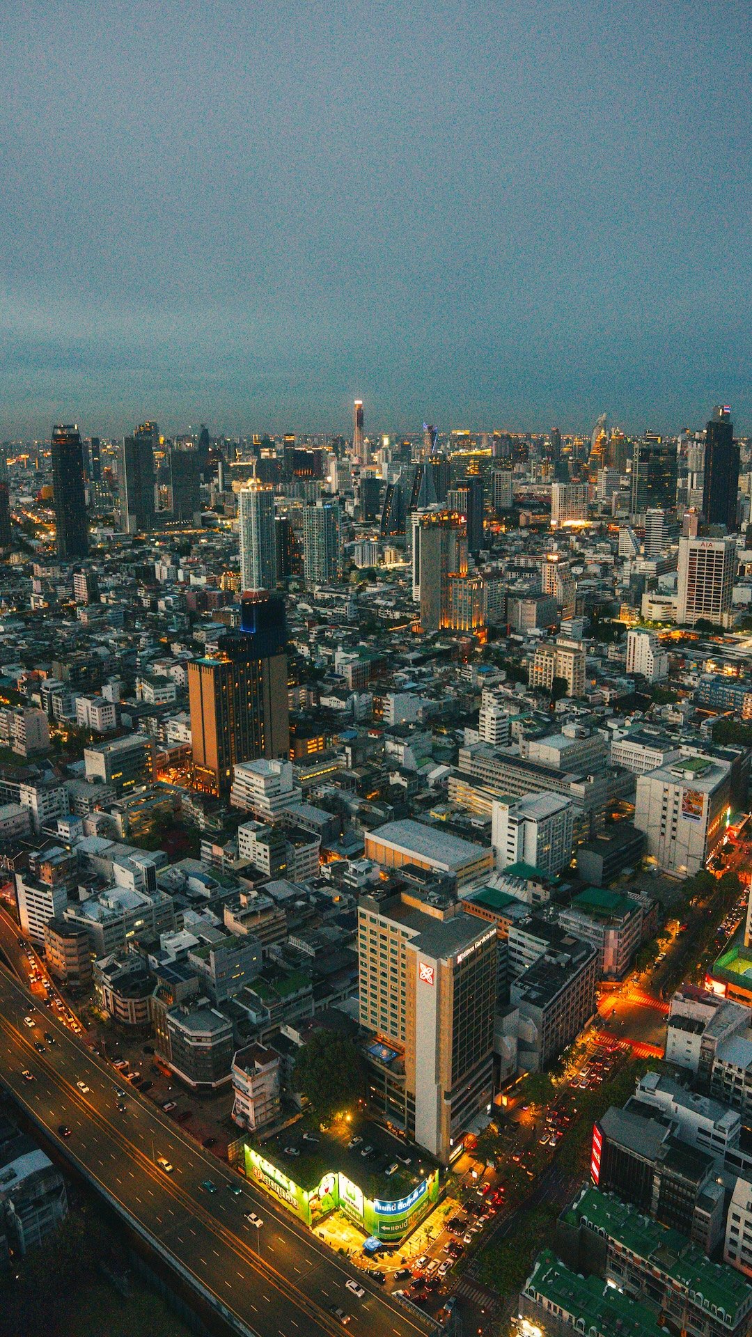 Uitzicht over een drukke stad met hoge gebouwen bij schemering, verlichte straten en wolken boven de skyline.