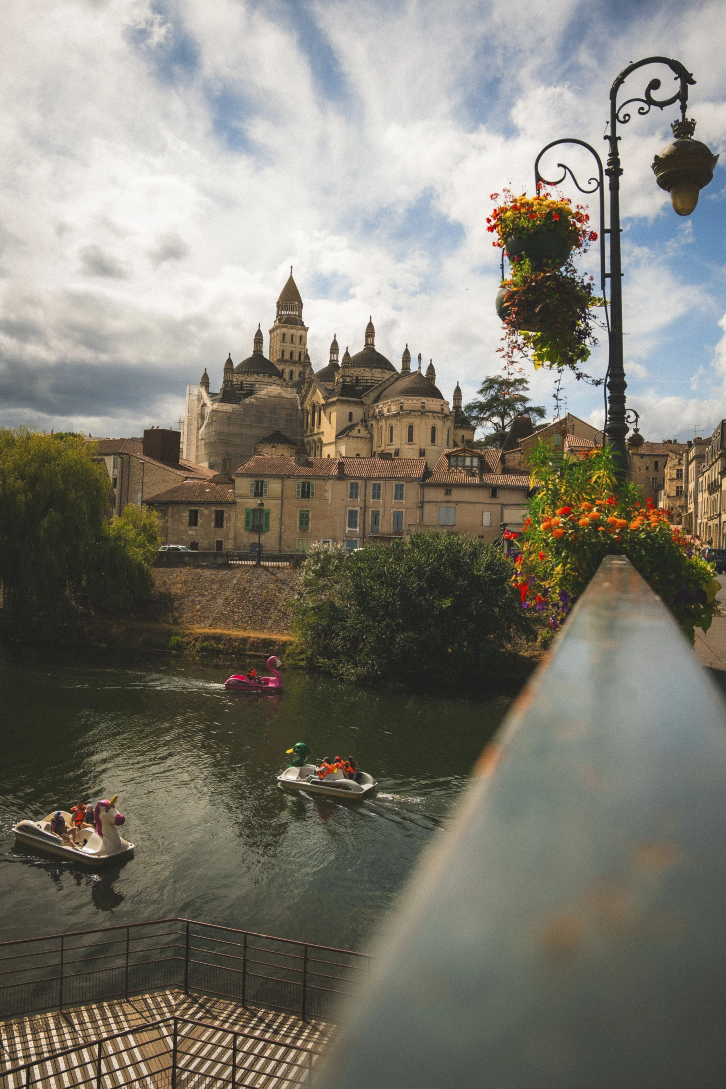 Uitzicht op een rivier met mensen die in dierenvormige zwembaden varen, naast een muur met bloemen en een oude kathedraal op de achtergrond onder een bewolkte hemel.