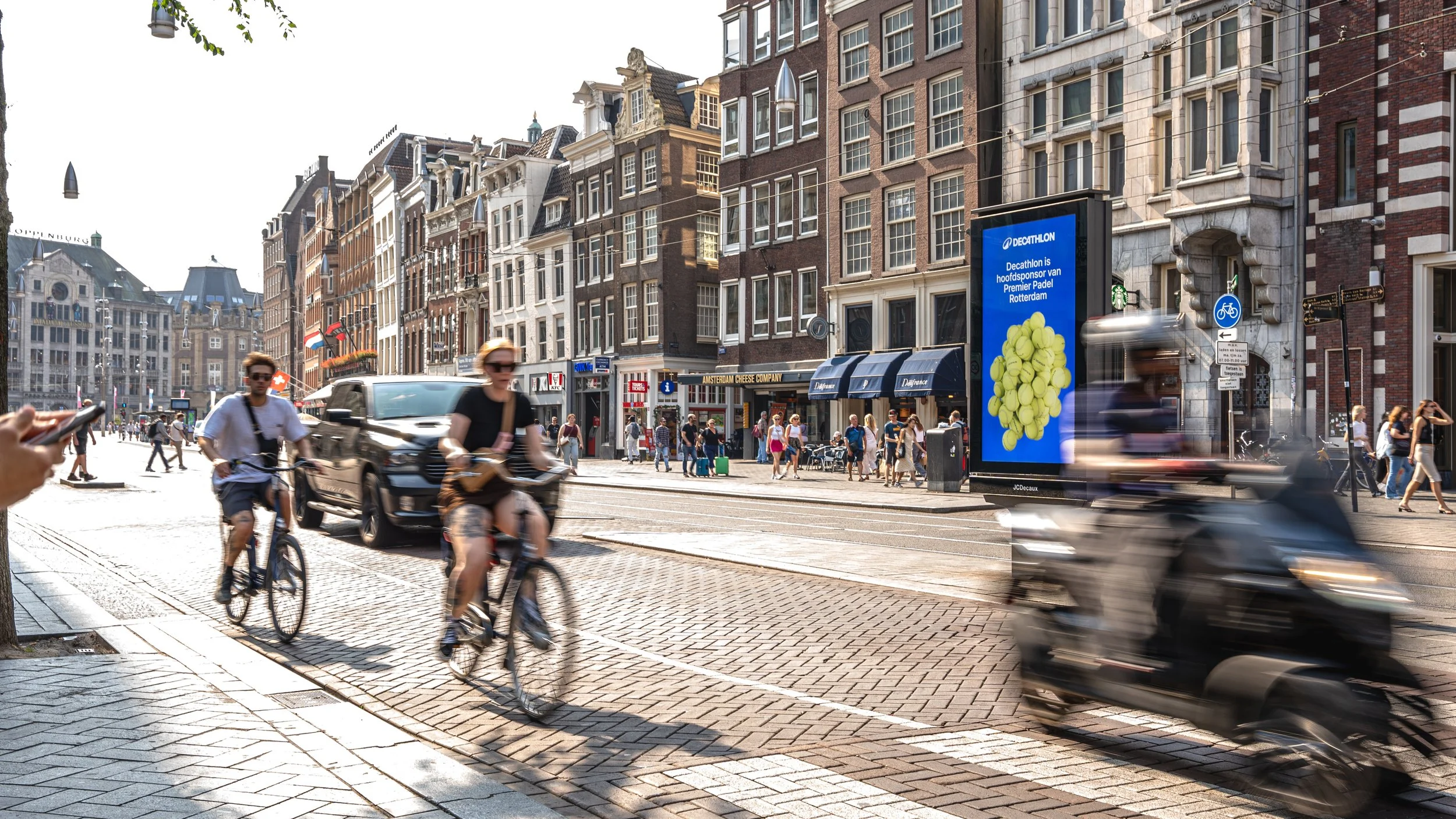 Straatbeeld van een drukke Amsterdamse straat met fietsers, voetgangers en voertuigen voor historische gebouwen en winkels.