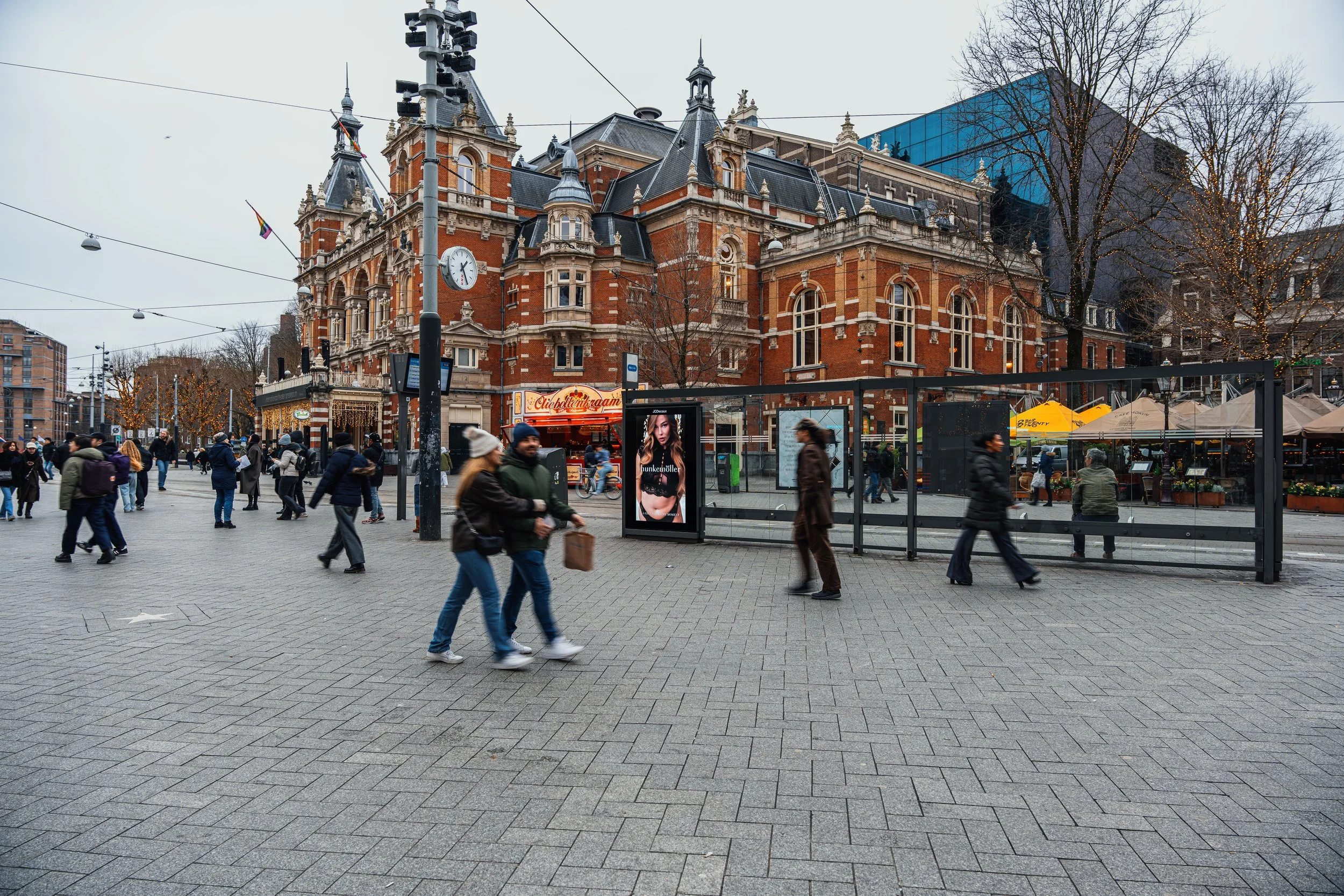 Stedelijke scène met mensen op een overdekte bus- of tramhalte voor een groot, oude, roodstenen gebouw met torens en een klok, omgeven door bomen en straatverlichting.