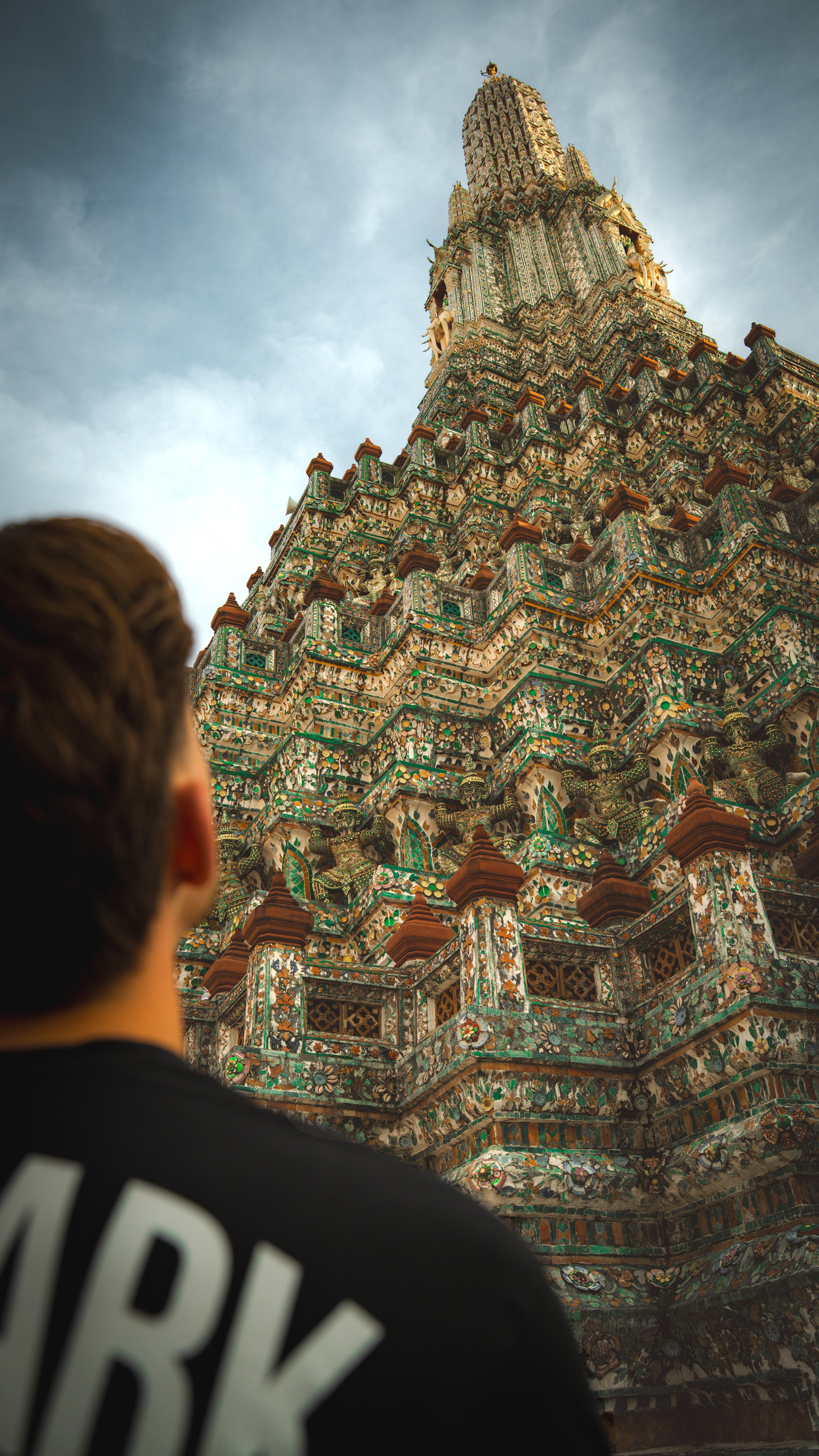Een persoon in de voorgrond kijkt naar een hoge, decoratieve tempel met veelkleurige keramische tegels en ornamenten, waarschijnlijk in Thailand.