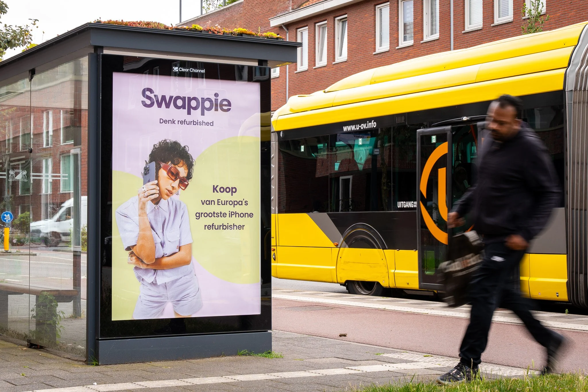 Een bushalte met een advertentie voor Swappie, een iPhone refurbisher, naast een gele en zwarte bus en een man die snel voorbijloopt.