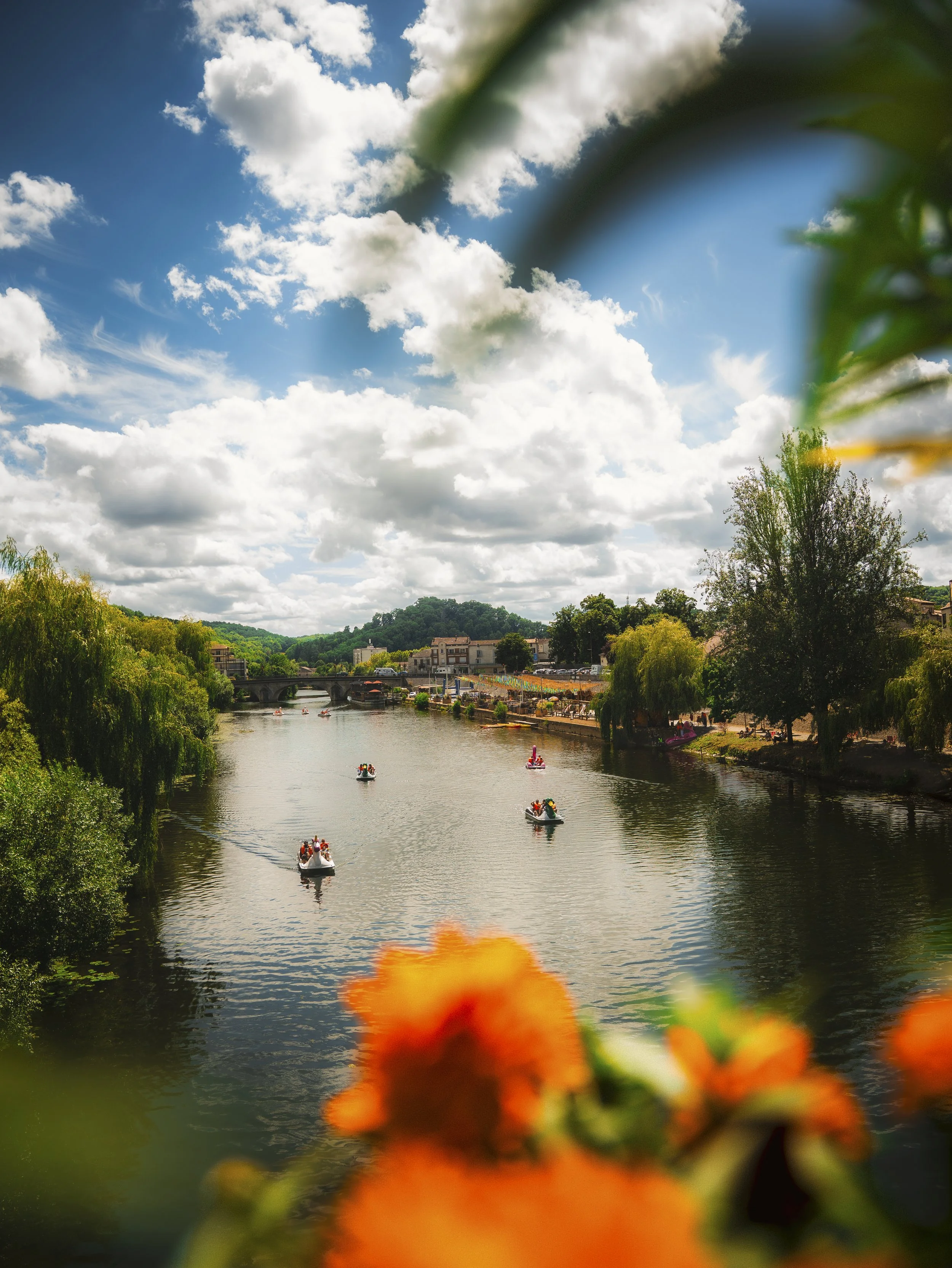 Een rivier met roeiboten, omliggende bomen, een brug en gebouwen in de achtergrond, met deels groene bladeren en oranje bloemen vooraan en een bewolkte blauwe lucht erboven.