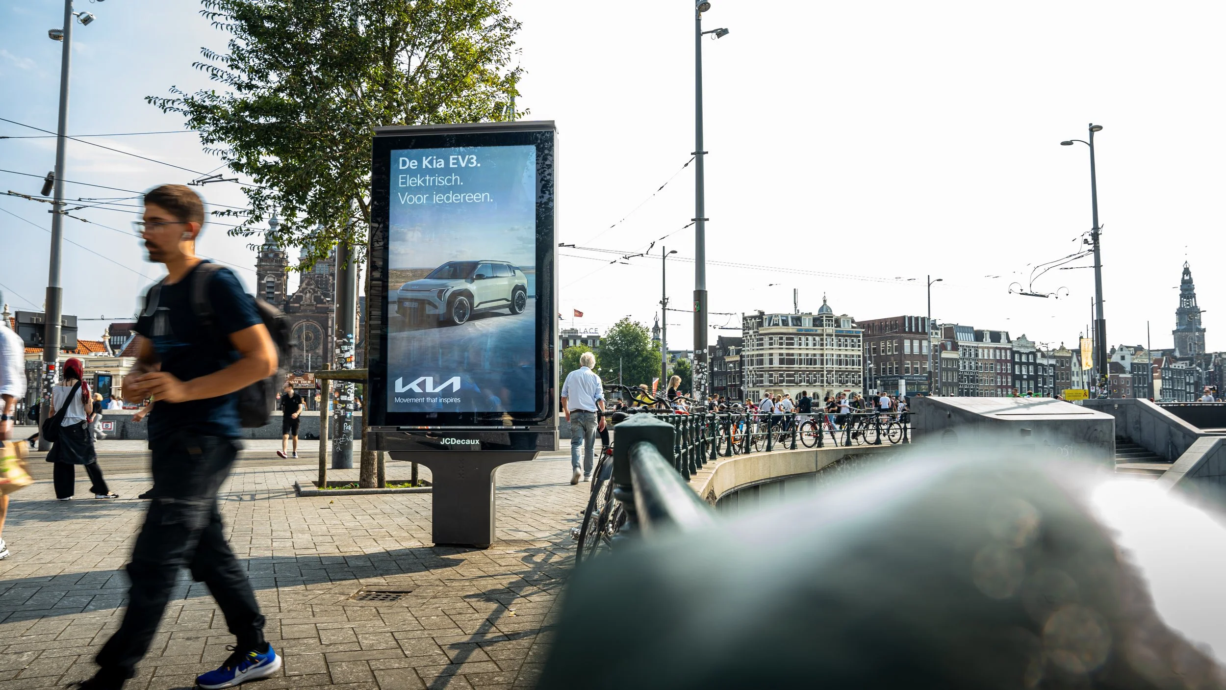 Stadscliché met mensen op het plein, een elektronische reclamebord dat een Kia EV3 auto adverteert, en historische gebouwen op de achtergrond, met een goed verlichte dag en een heldere lucht.