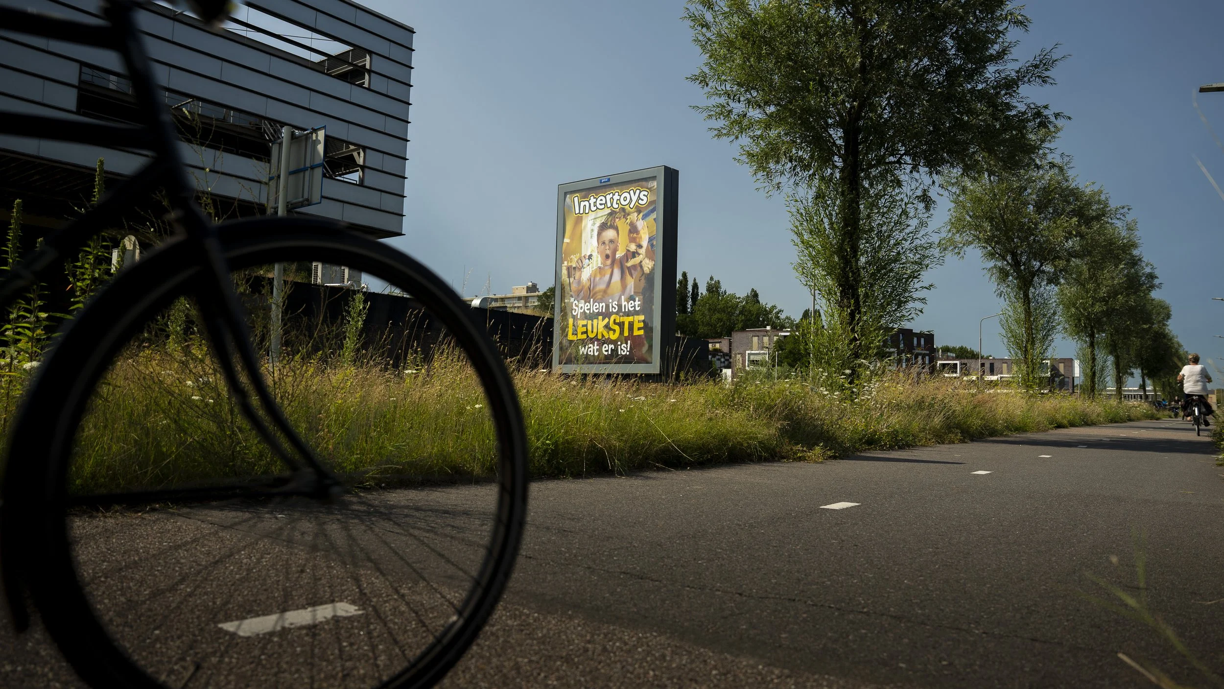 Bord met een reclame voor Intertoys met de tekst 'Spelen is het leukste wat er is!', een fietser rechts op de achtergrond, en een fietspad met witte lijnen.