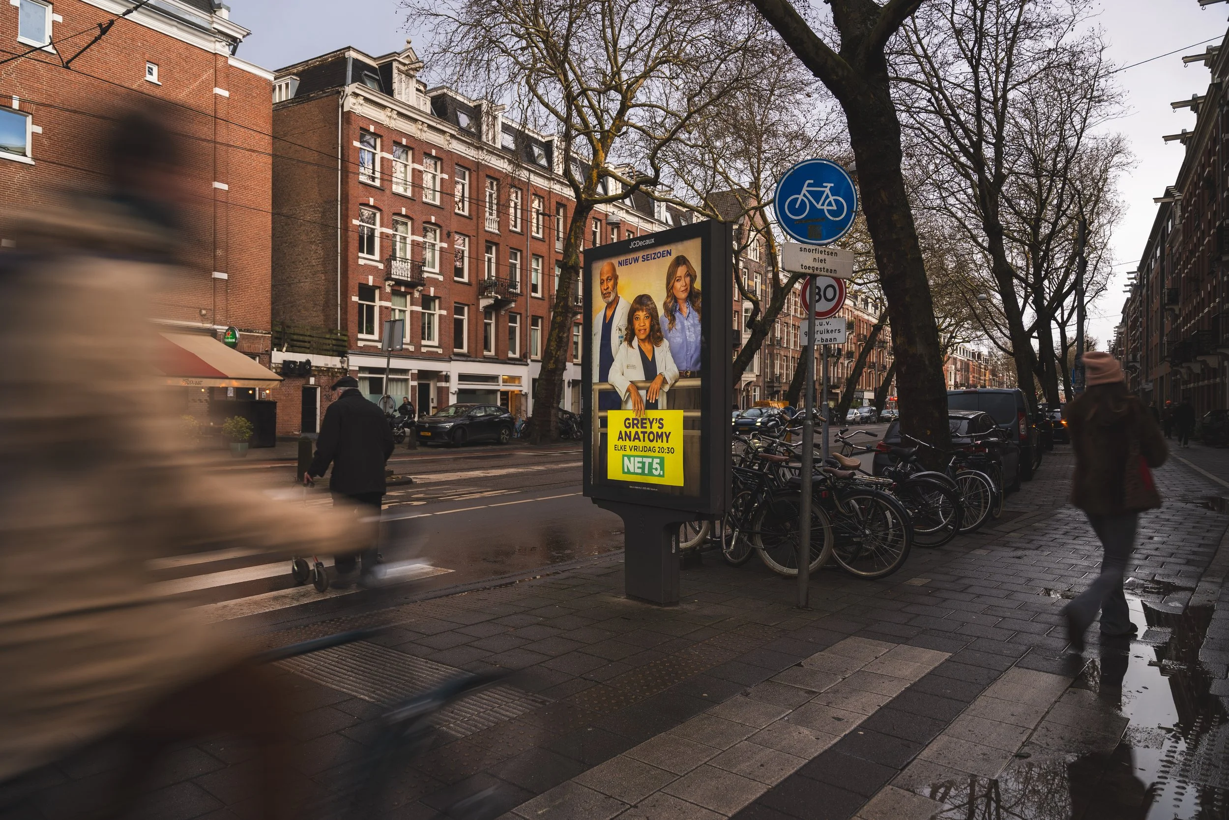 Stadsplein met fietsroutebord, reclamebord voor Grey's Anatomy op scherm, gekleurde bakfietsen geparkeerd, mensen die over straat lopen, rijen oude stenen gebouwen, bomen, auto’s en regenplassen op straat.