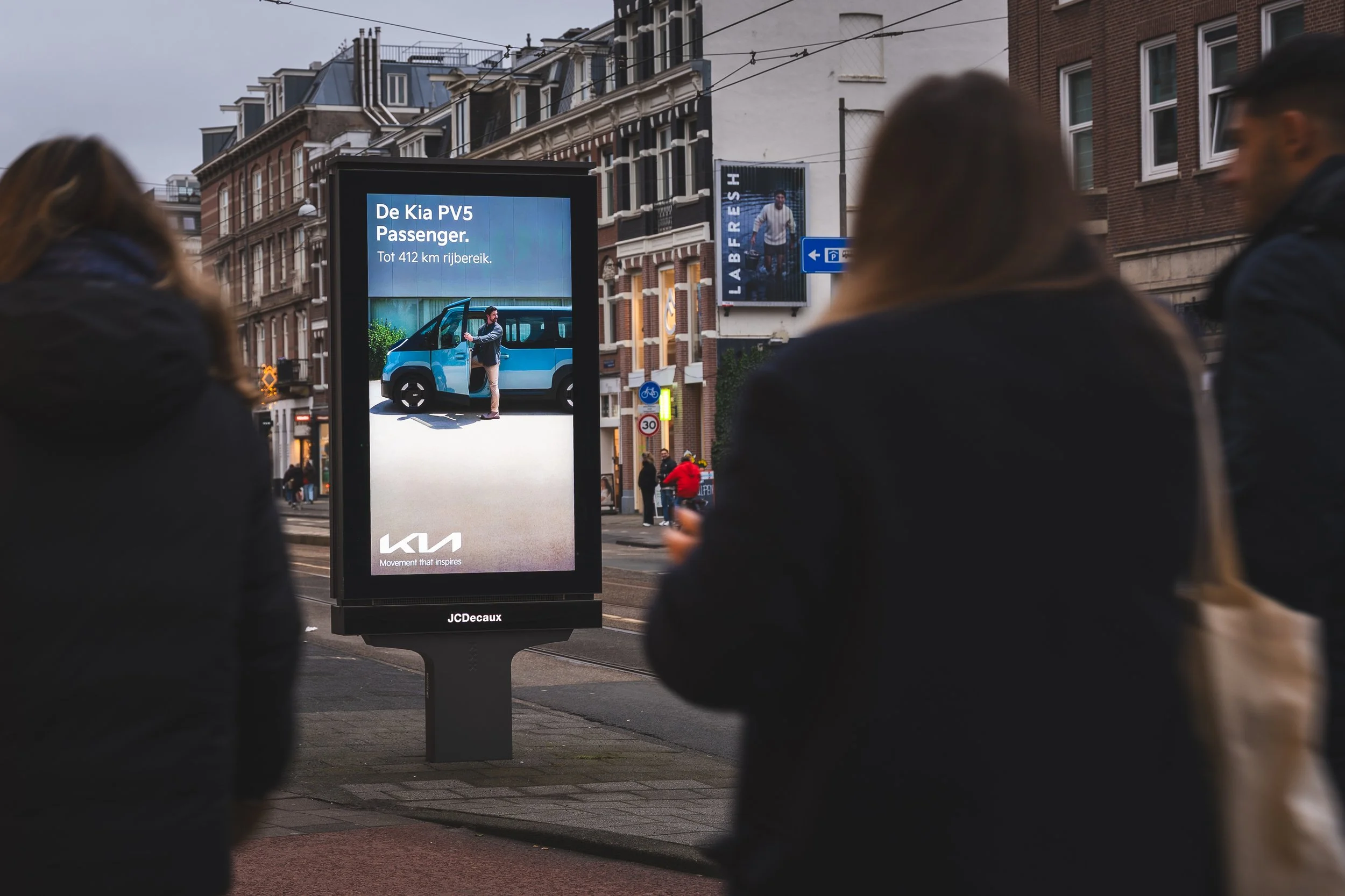 Drie mensen staan op straat bij een digitale reclamezuil, met gebouwen en straatverkeer op de achtergrond. Op het scherm staat een advertentie voor een Kia PV5 Passagier auto.
