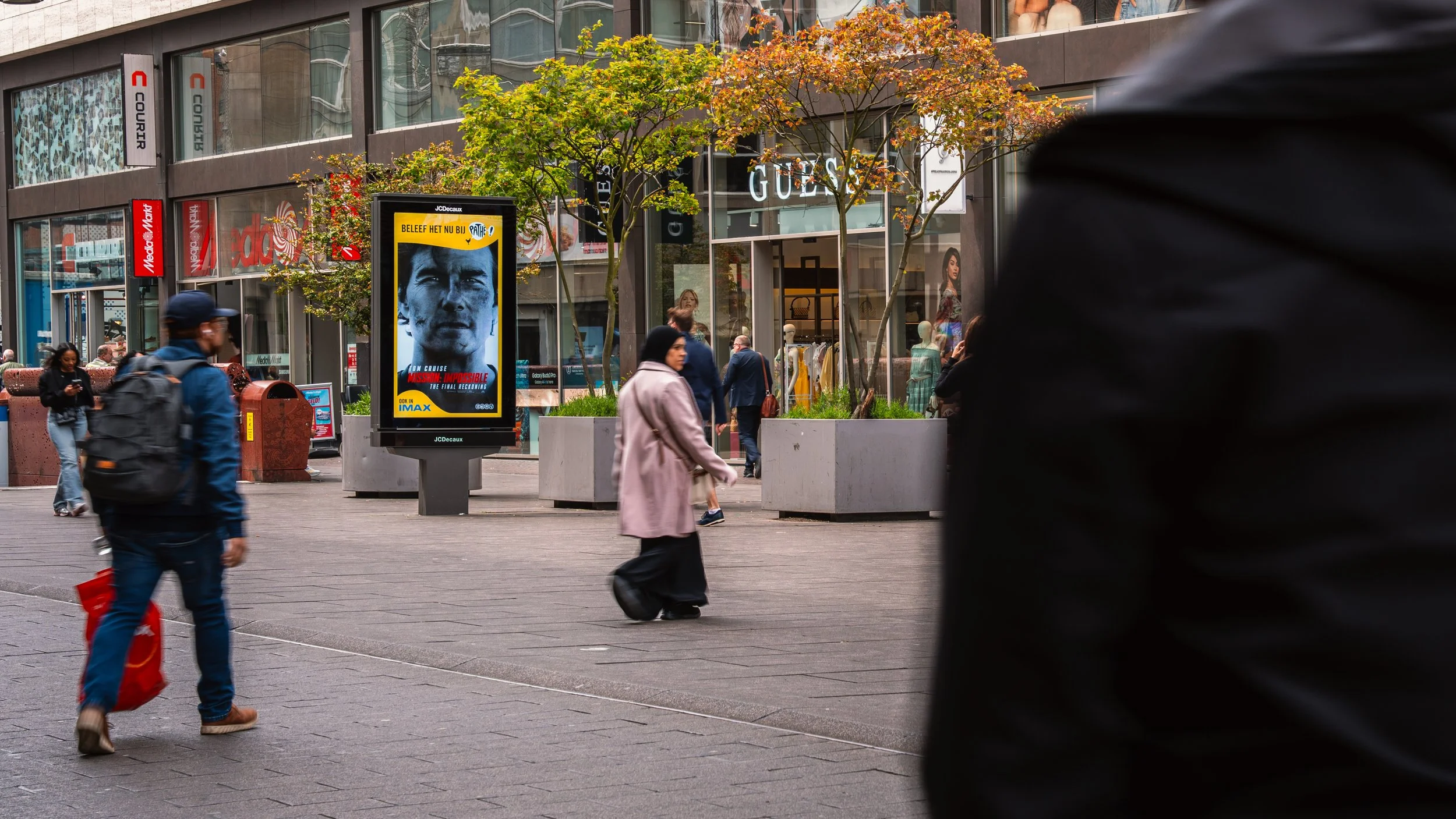 Stedelijk winkelcentrum met wandelaars, winkels, bomen en een digitale reclamezuil.