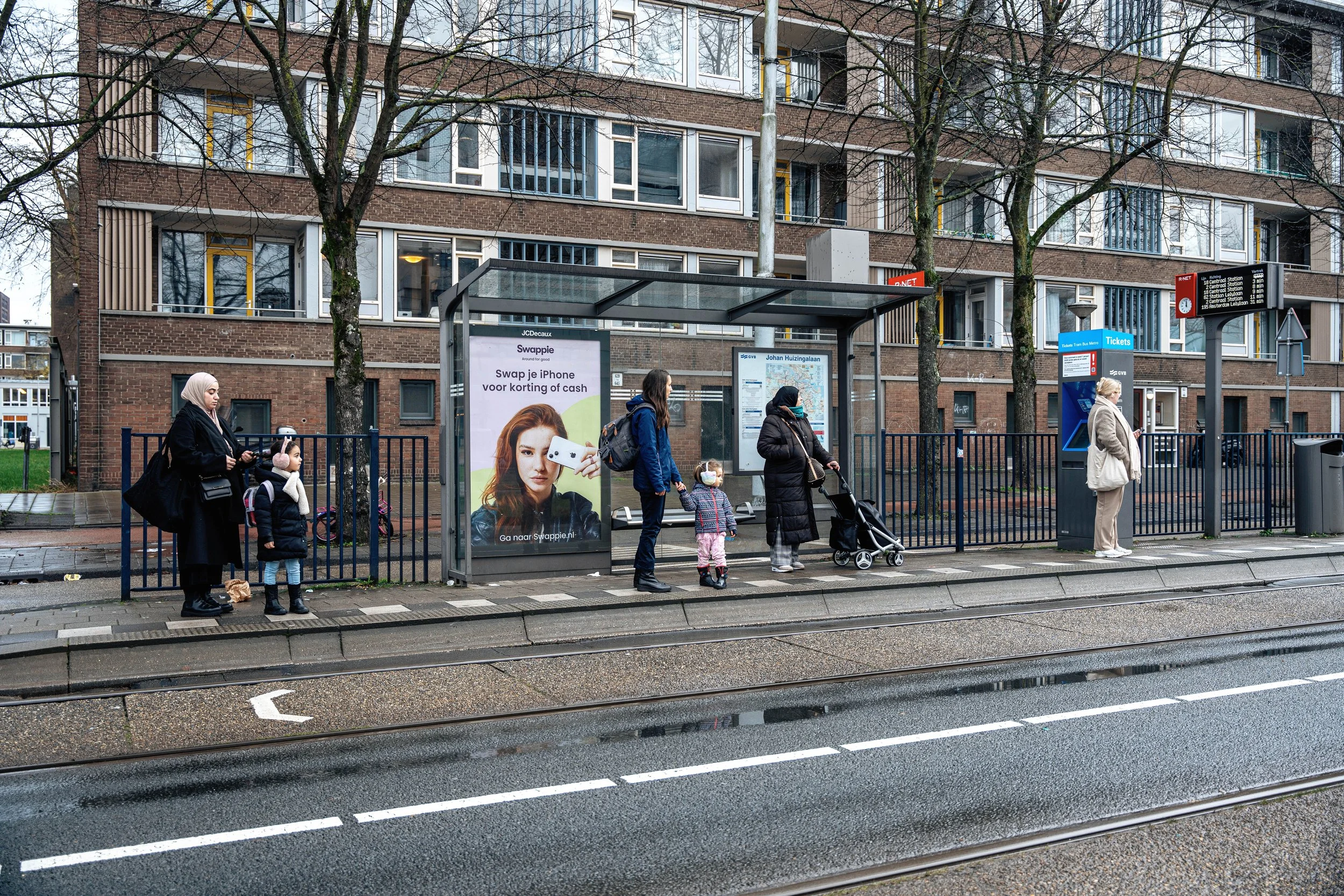Mensen wachten in de regen bij een bushalte in een stadswijk, met een grote appartementsgebouw op de achtergrond.