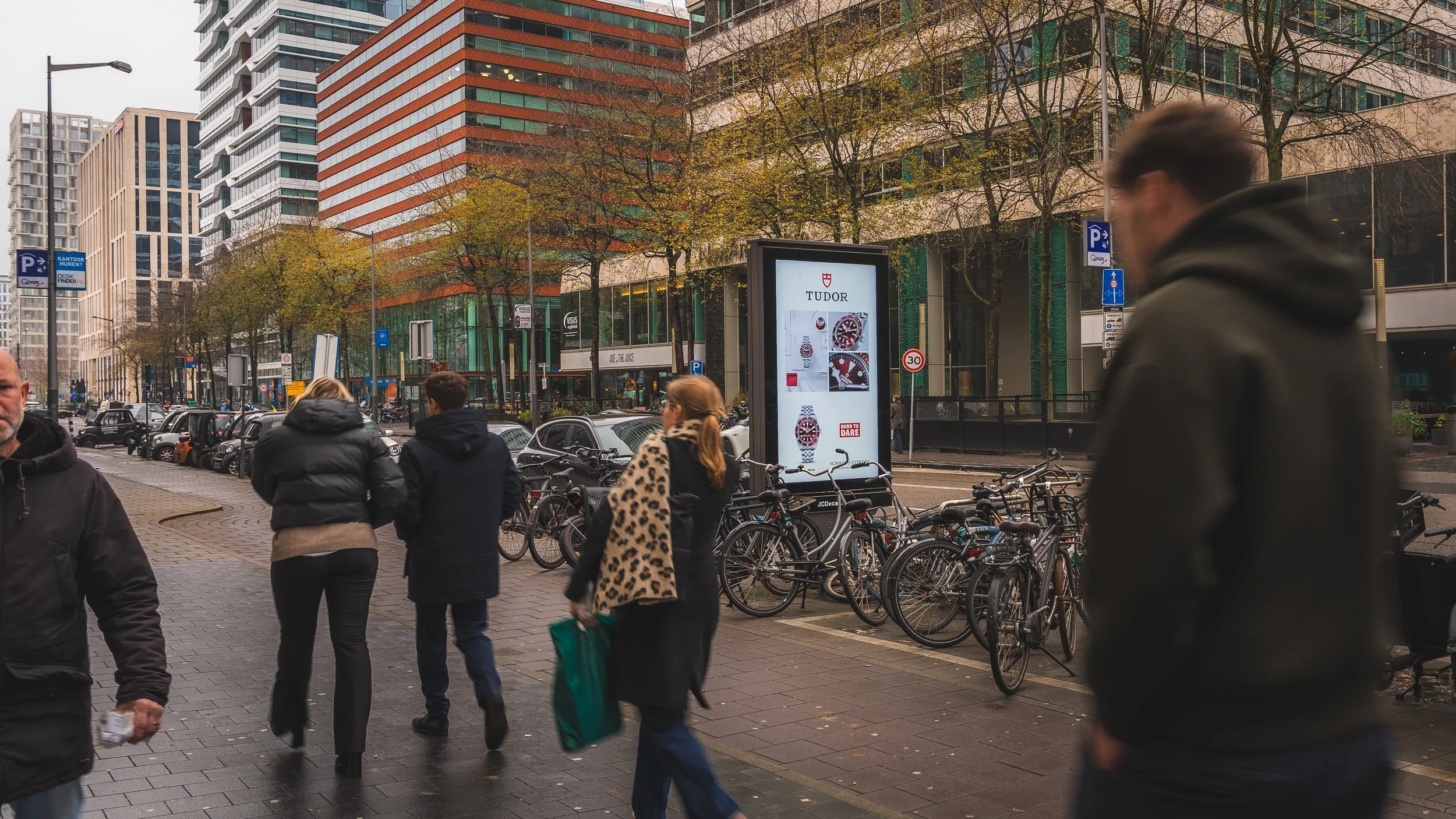 Straat met voetgangers, fietsen en moderne gebouwen met wandelende mensen op een bewolkte dag in een stedelijke omgeving.
