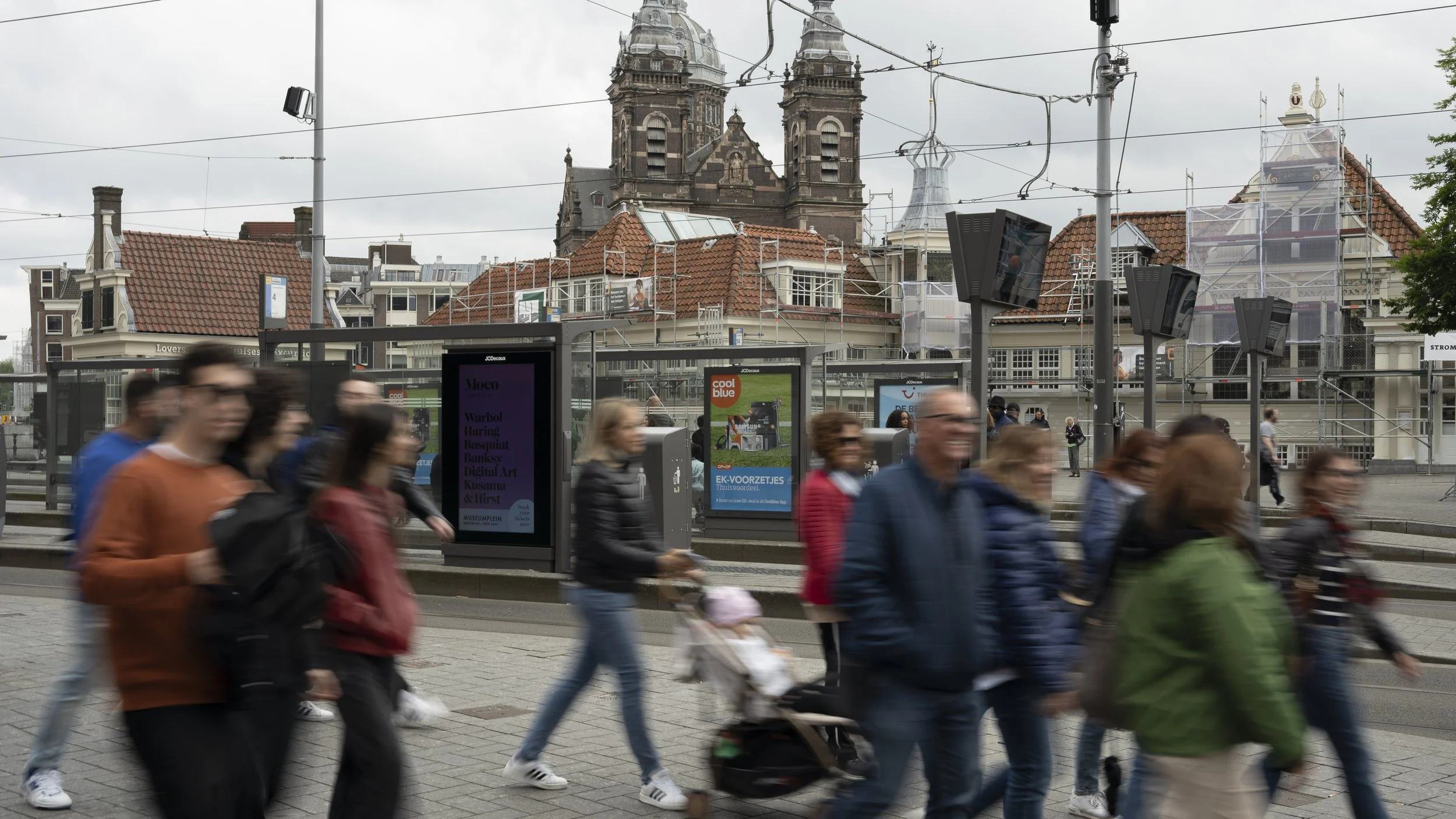 Mensen lopen over een stadsplein met trams en een historische kerk op de achtergrond, onder een bewolkte hemel.