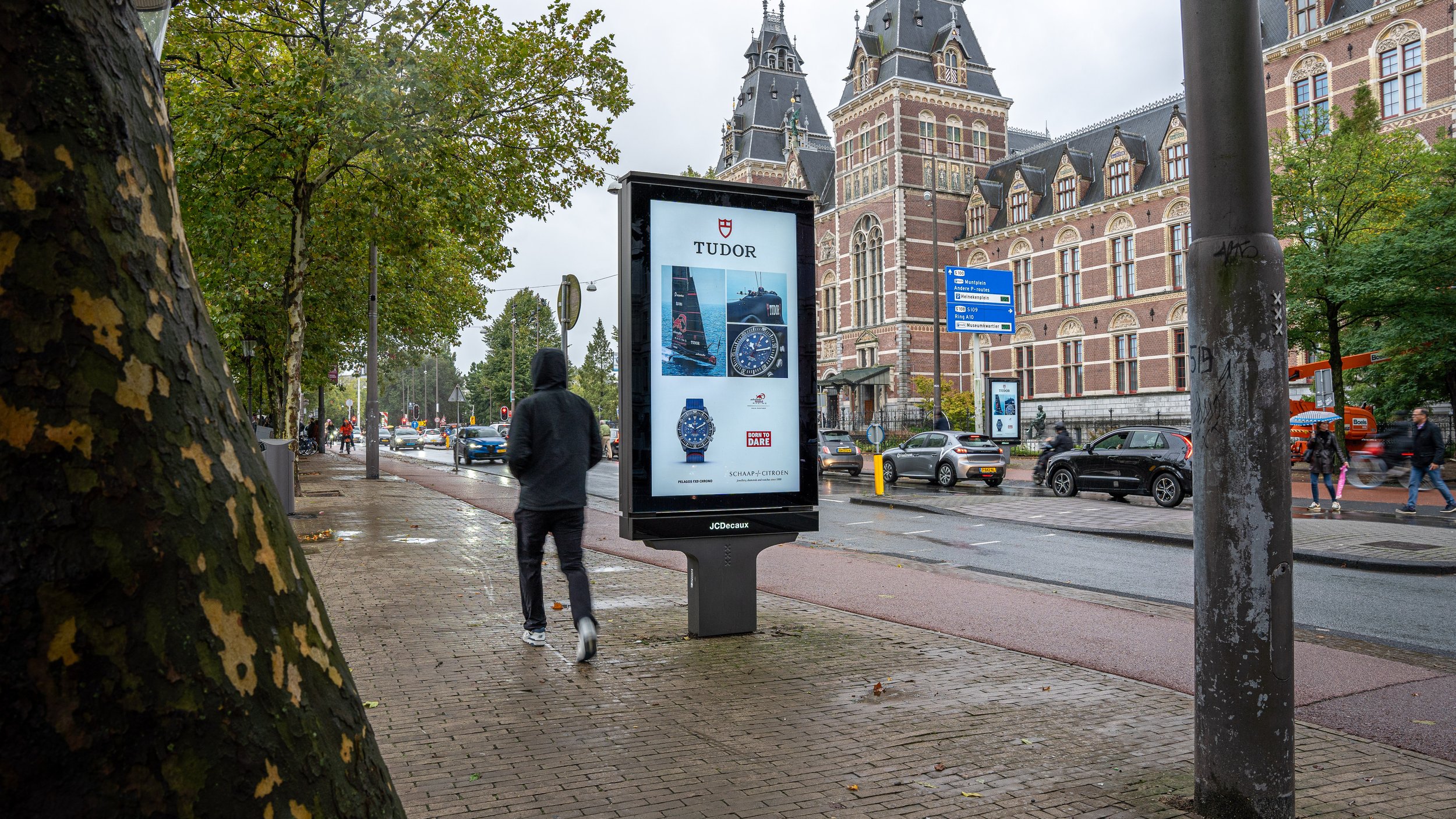 Stadsscene in regenachtig weer met voetgangers, auto's en een reclamezuil voor horloges voor Tudor in de stad met een groot gotisch gebouw op de achtergrond.