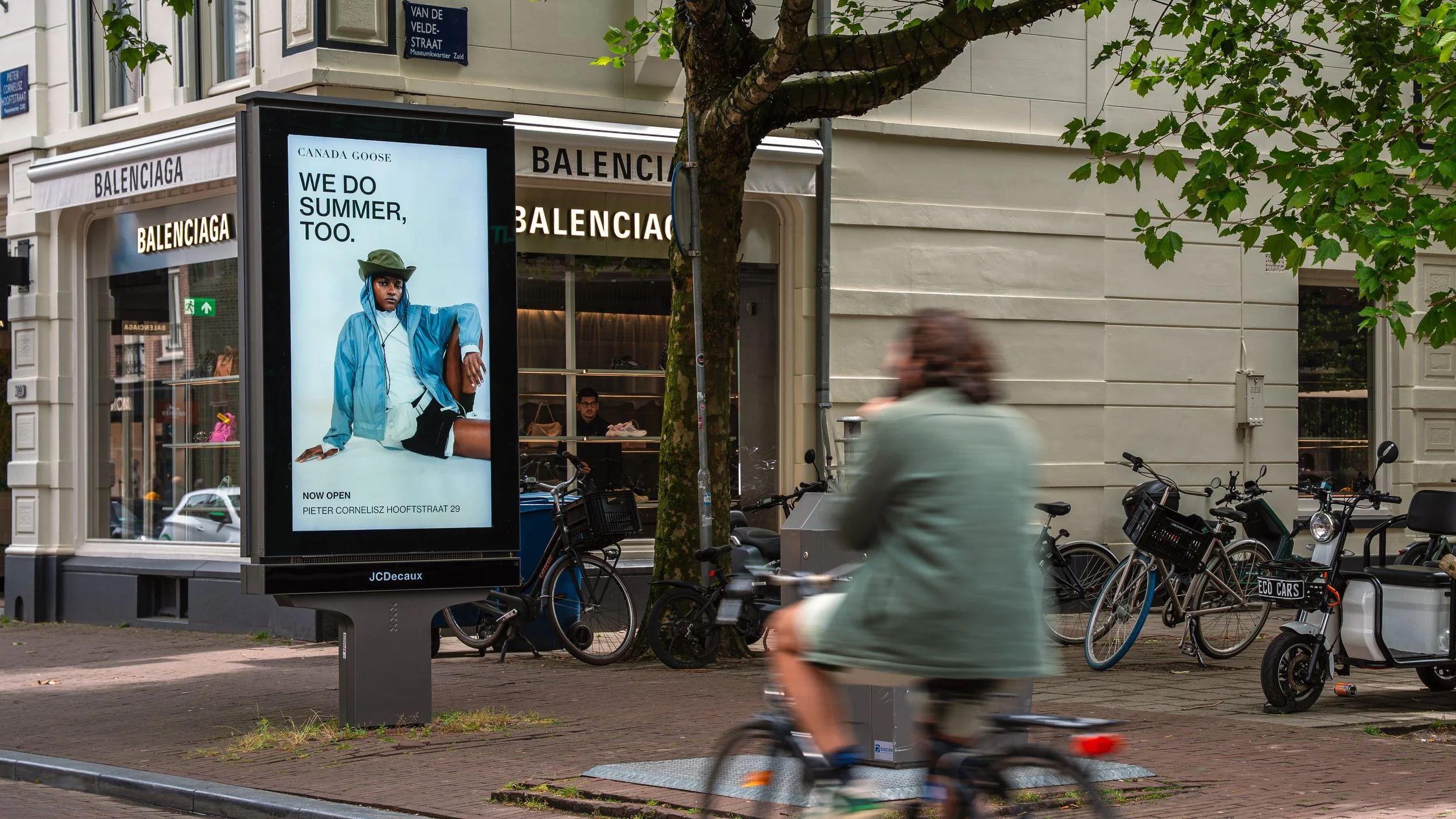 Digital billboard on a city street advertising a store opening with a model in casual clothing and a hat. Surrounding buildings have store signs and several bicycles are parked along the sidewalk, with one person riding a bike past.