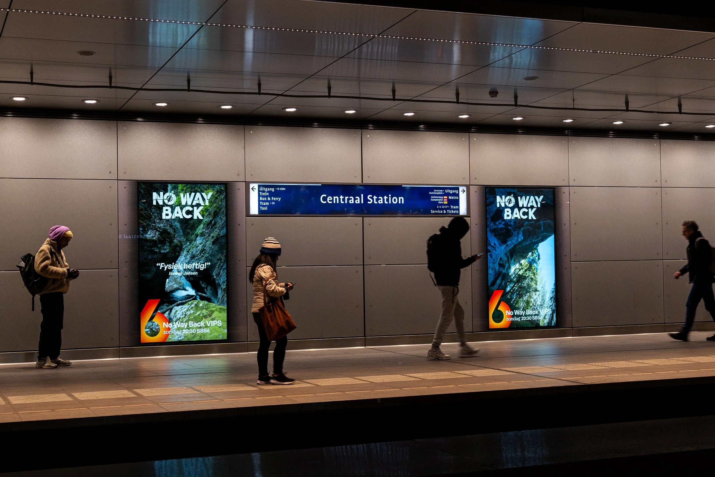 Mensen wachten op de metro bij centraal station, met digitale bewegwijzering en reclameborden op de achtergrond.