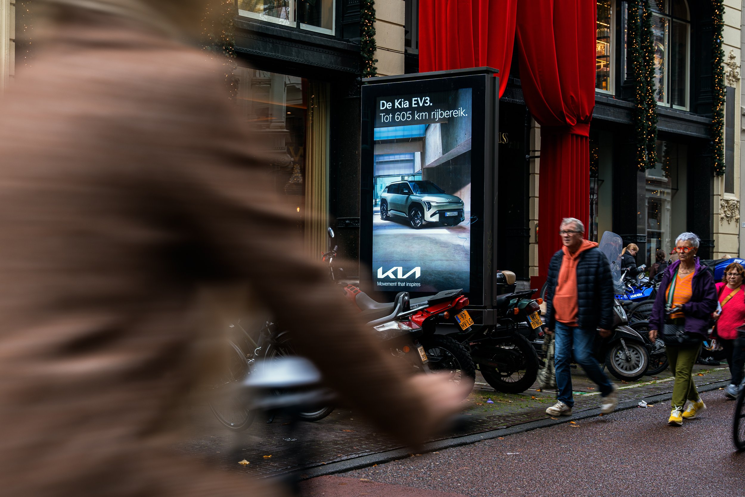 Een straatmet grote digitale reclameborden die auto's en motorfietsen promoten, met mensen die voorbij lopen op een regenachtige dag.