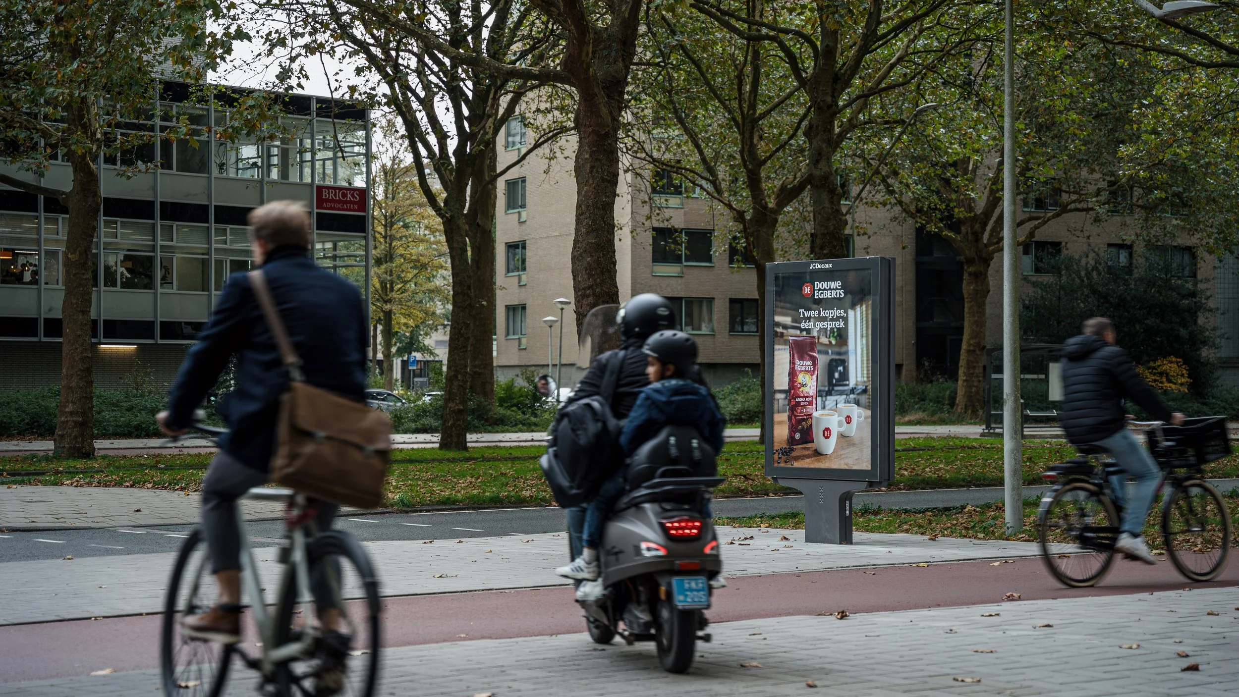 Drie mensen fietsen en een persoon rijdt op een scooter op een stadsfietsroute, met bomen, huizen en een digitale billboard op de achtergrond.