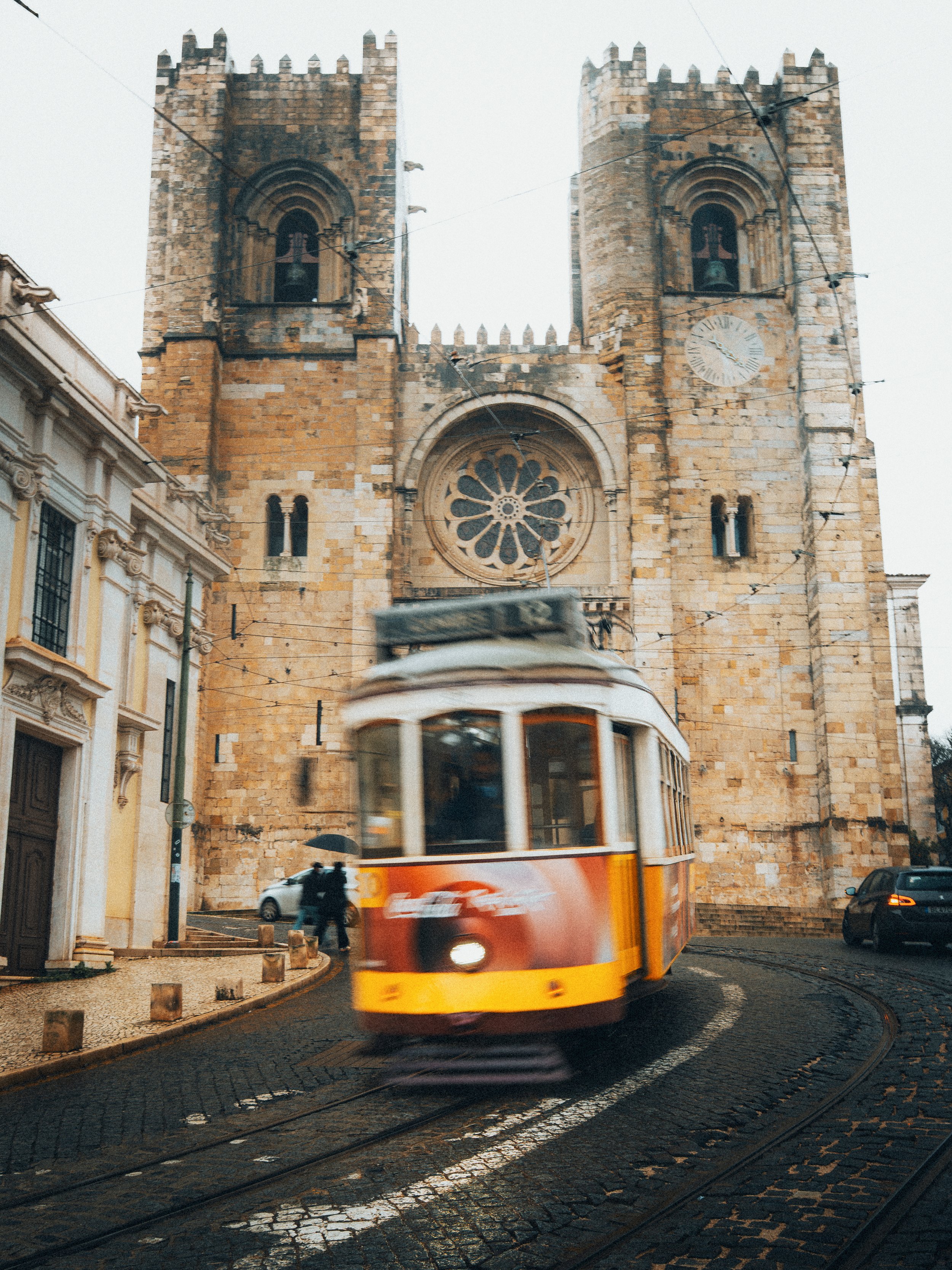 Een gestileerde foto van een gele en rode tram die door een straat rijdt voor een oude kathedraal met twee torens en een roosvenster.
