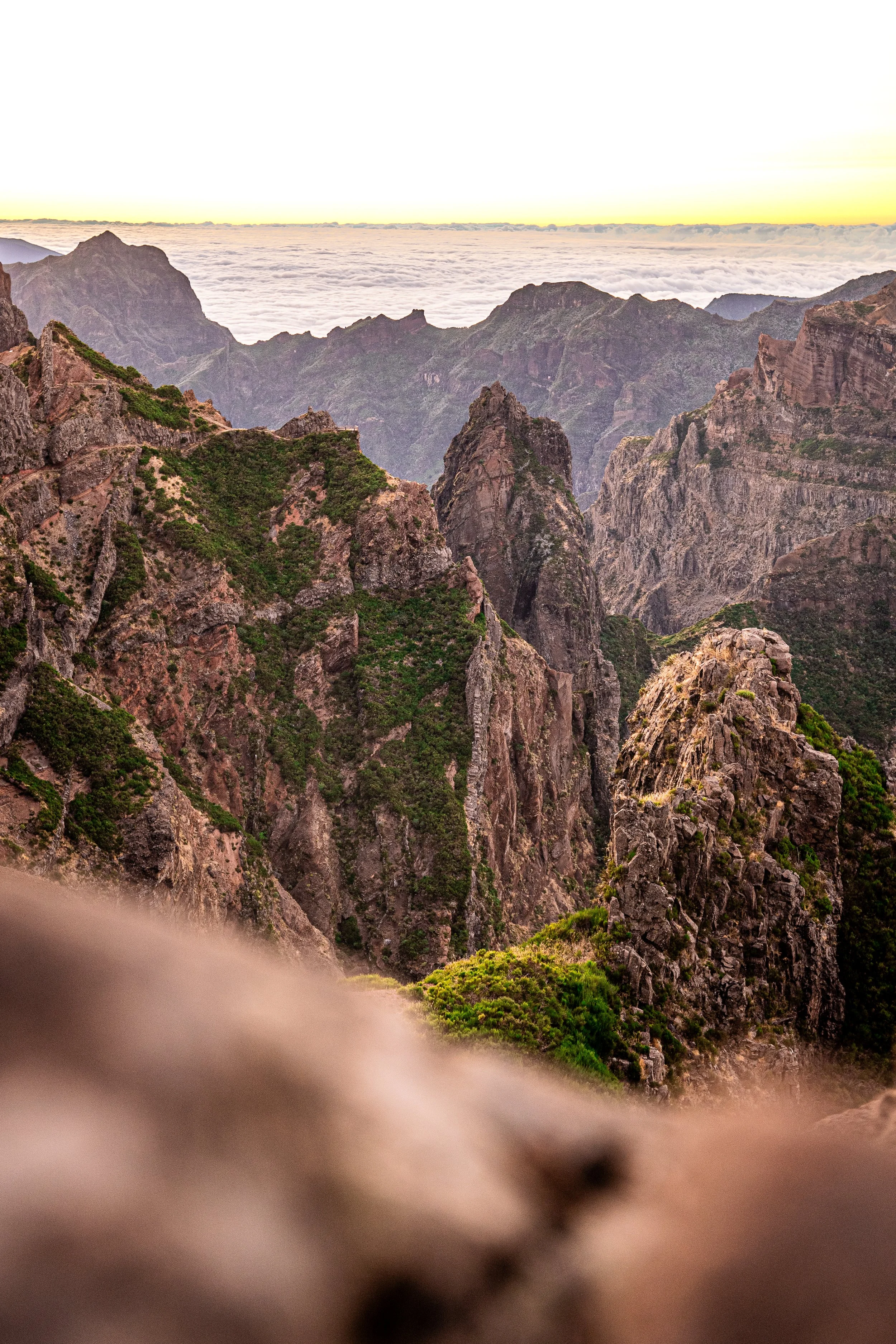 Een berglandschap met rotsachtige bergen en groene vegetatie, uitzicht op een wolkenlaag en een heldere lucht tijdens zonsopgang of zonsondergang.